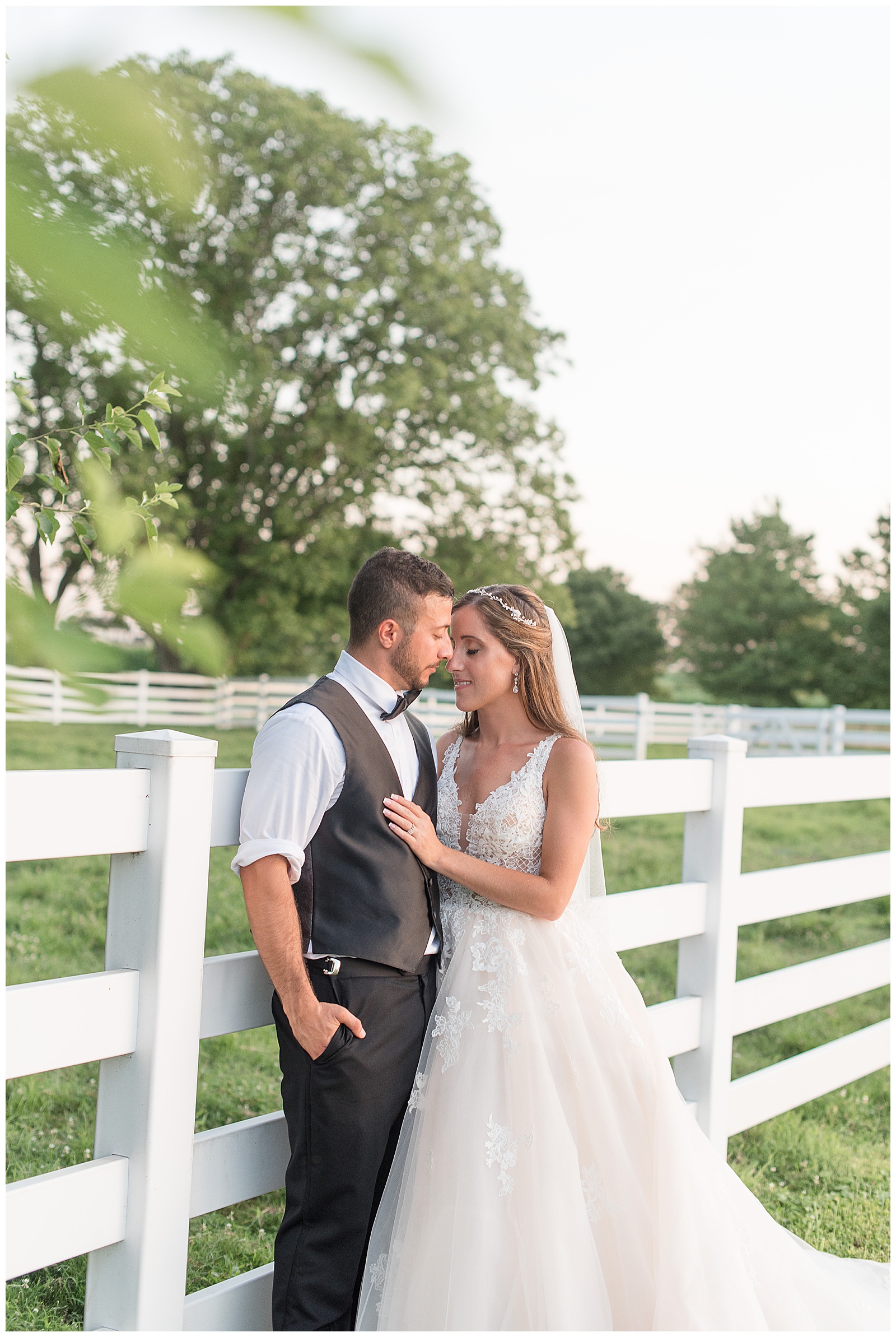 groom leans against white fence with hand in pocket as bride leans against him smiling at the silverstone barn