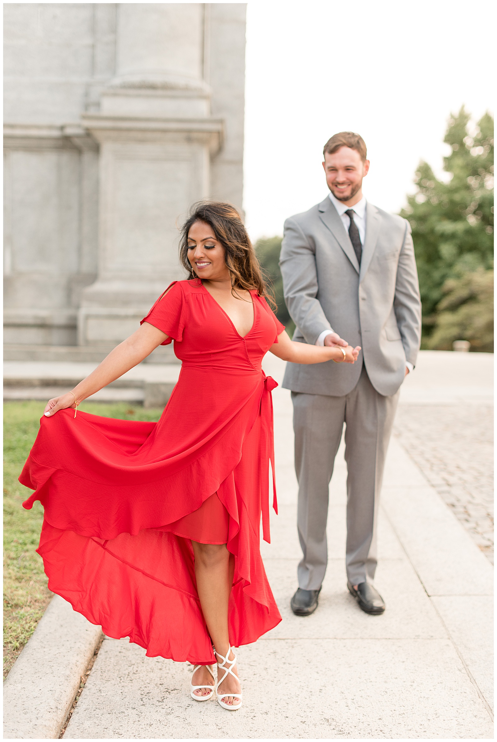 Girl in flowy red dress smiling down towards hand as guy holds her hand from behind smiling at her