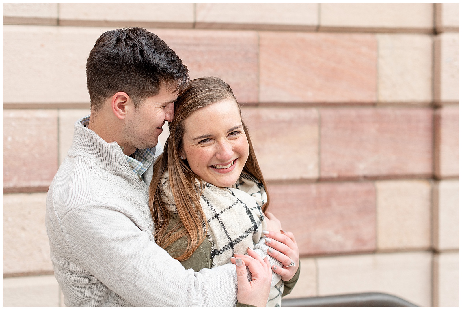 guy has arms wrapped around girl nuzzling into her temple as she is smiling at the camera