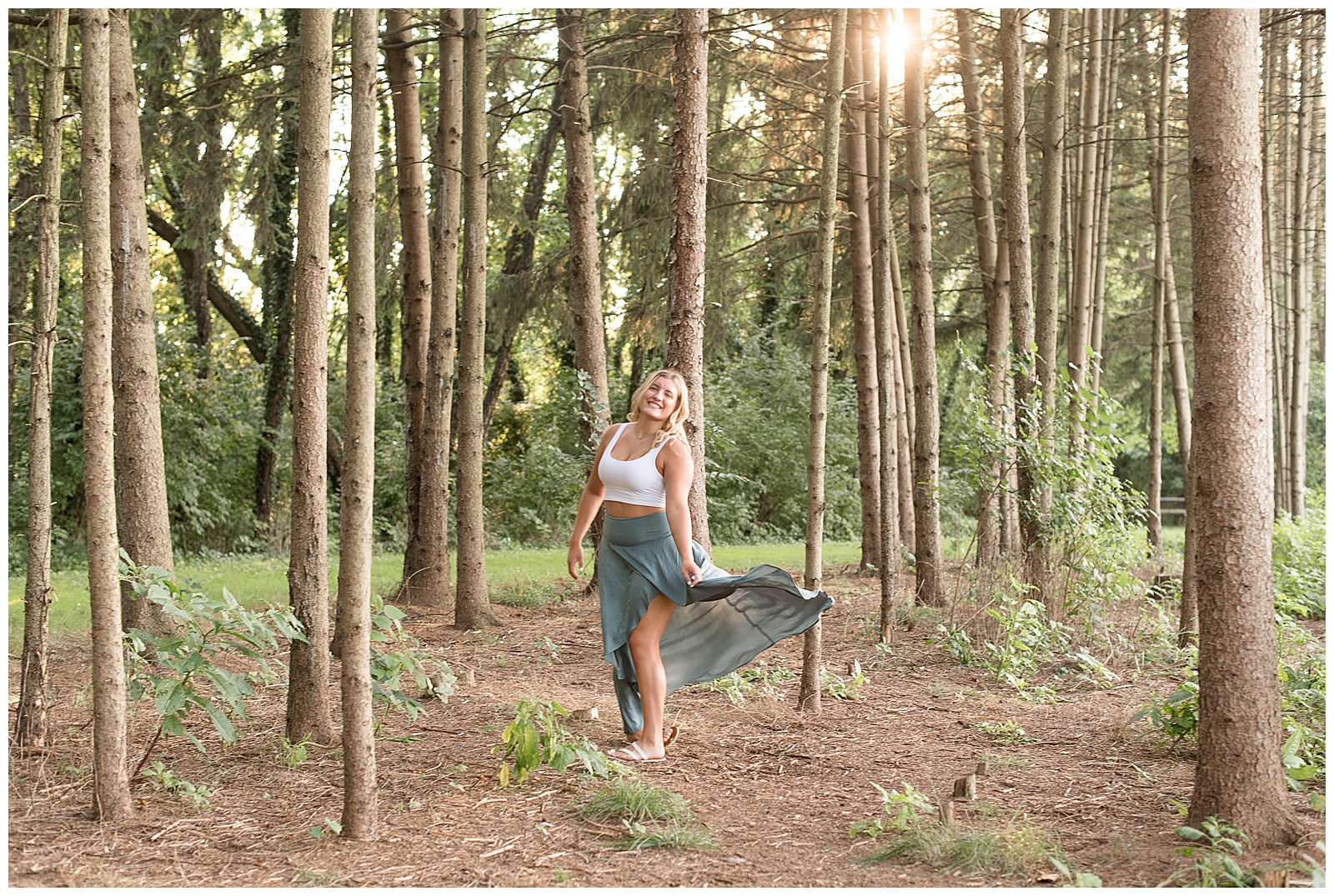 girl turned smiling with skirt blowing in wind among evergreen tree row at overlook park