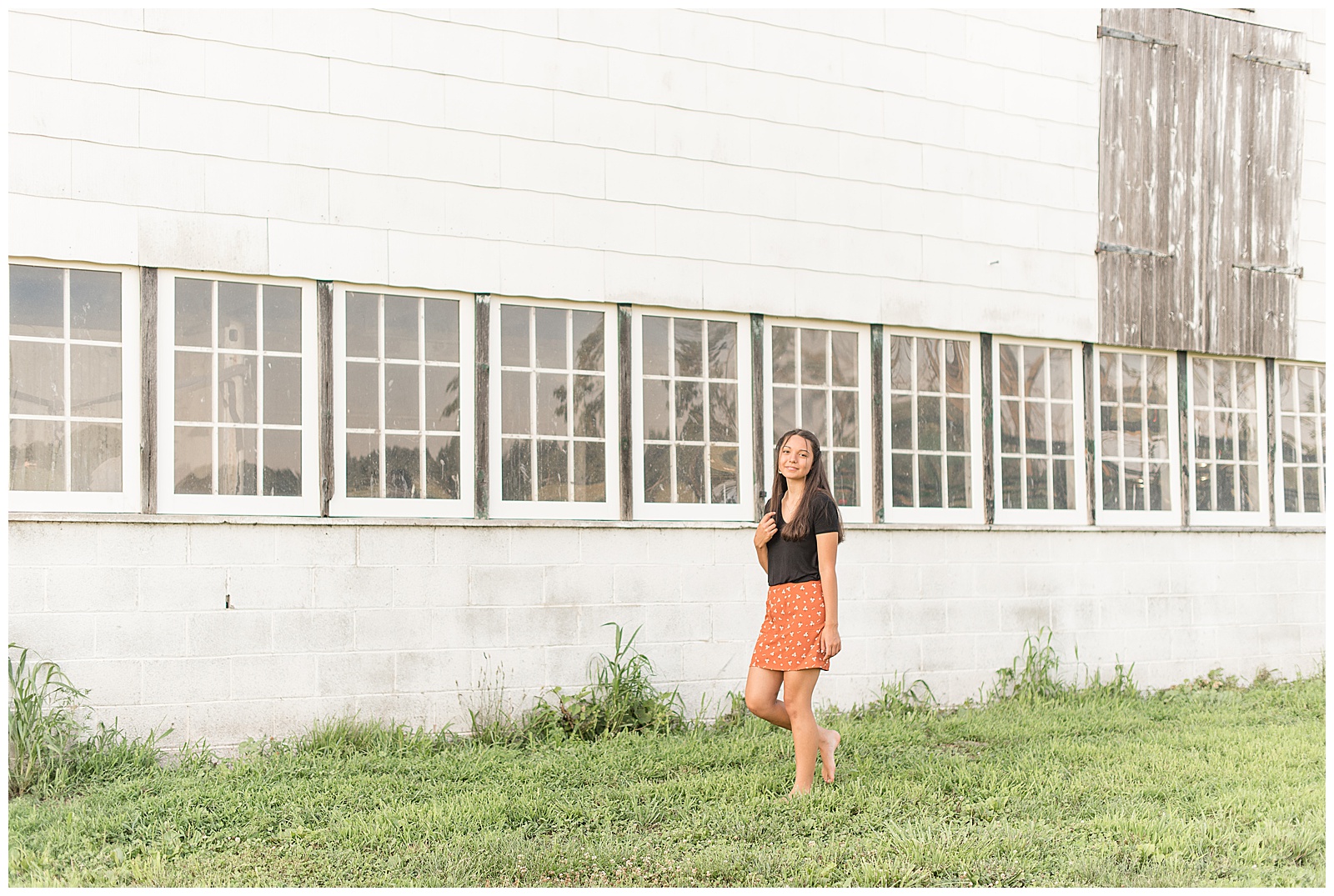 girl walking along rustic white barn in grass smiling at camera in lancaster pennsyvlania