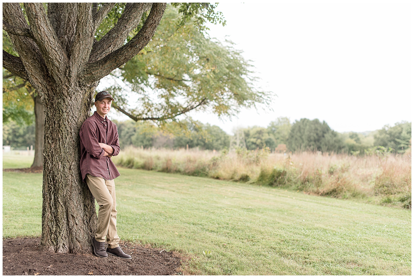 senior guy leaning against tree trunk arms folded wearing baseball hat in beautiful park