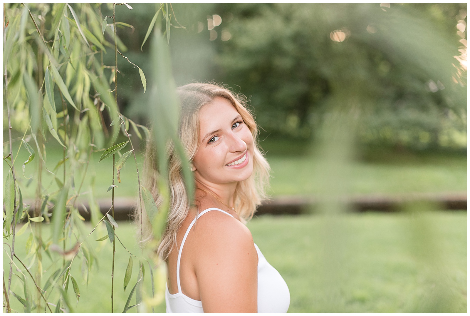 senior girl glancing over right shoulder smiling at camera behind willow tree in lancaster pennsylvania