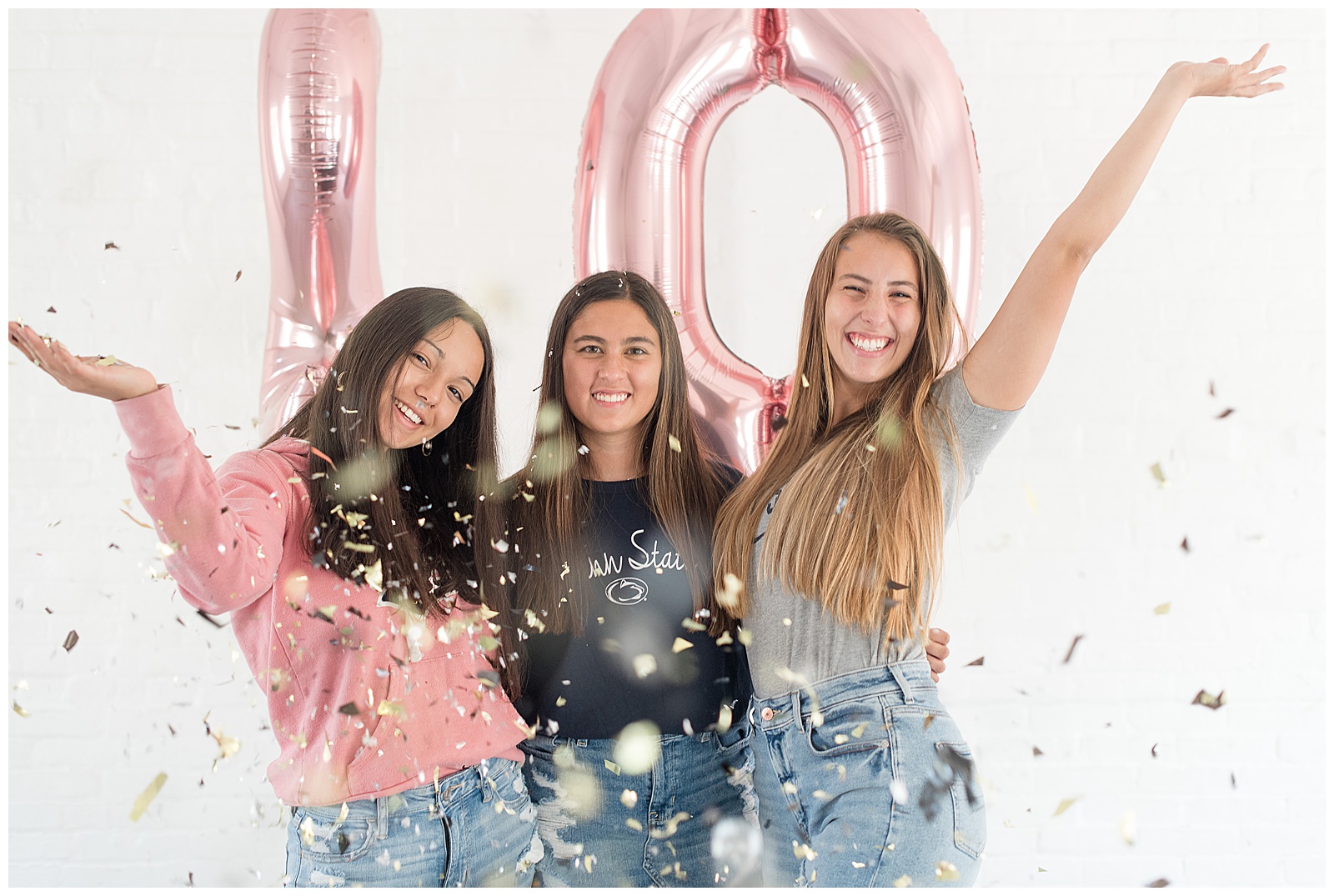 three senior girls hugging and smiling behind confetti along white wall with balloons at hingeworks