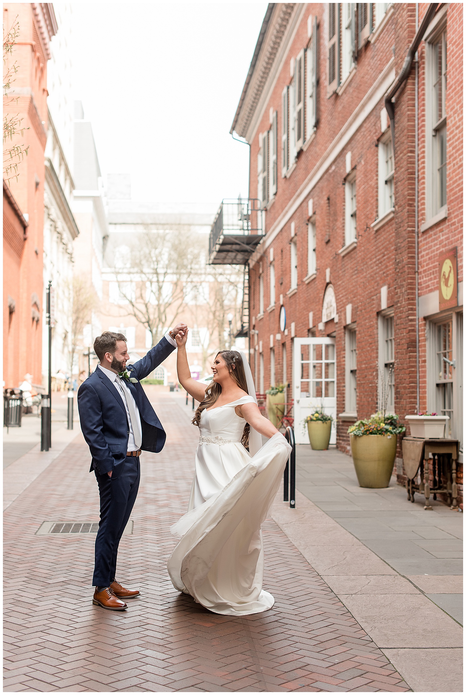 groom twirls bride as she flairs her bridal gown train on cobblestone street by central market