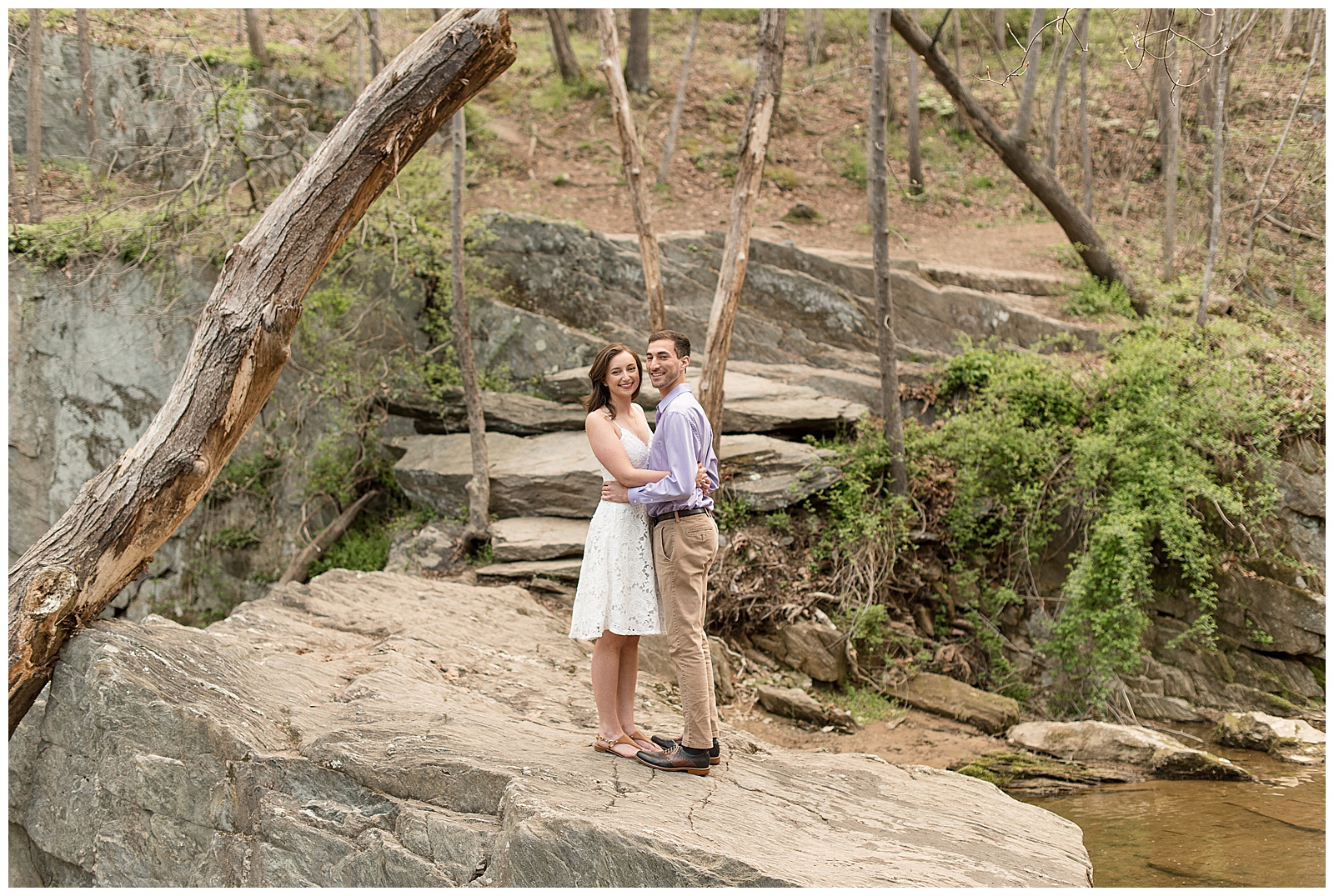 engaged couple hugging tightly on large rock in woods looking at camera in northern maryland