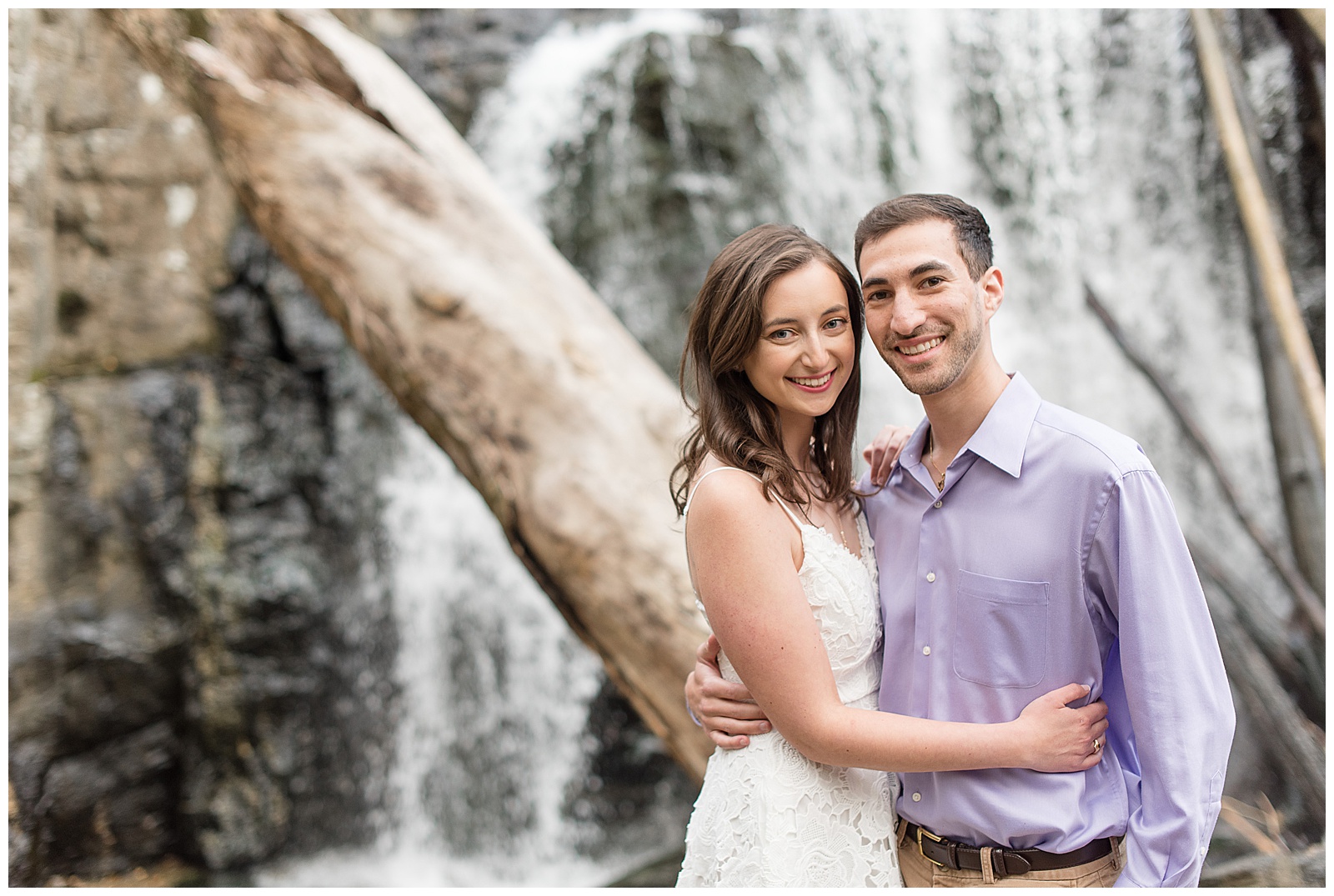 couple hugs smiling at the camera with kilgore falls and large downed tree behind them