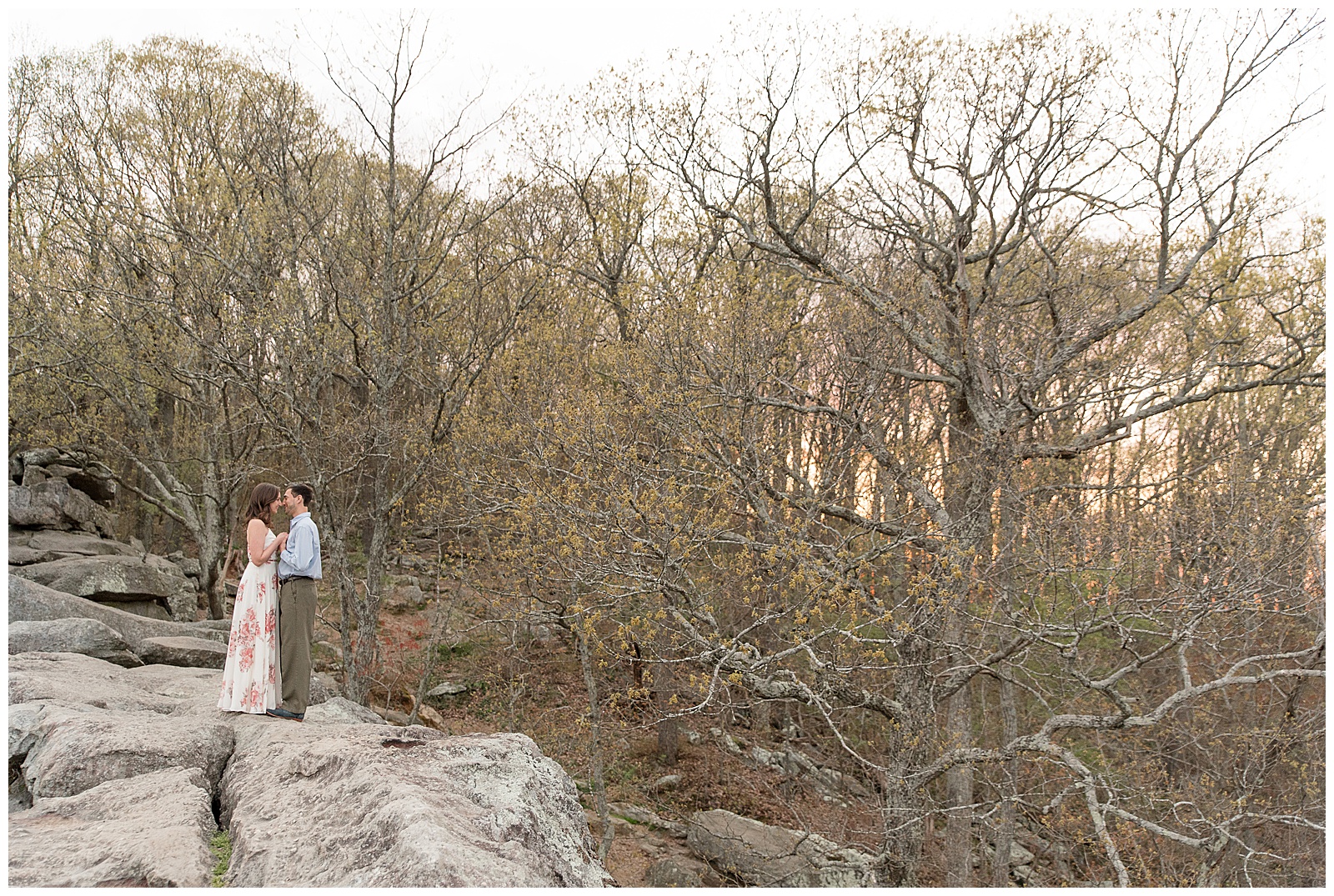 engaged couple standing close on large rock on left side of photo with forest behind them at rocks state park