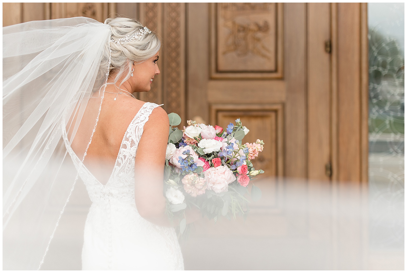 back of bride with long veil blowing as she glances over her right shoulder holding bouquet by wooden doors in maryland