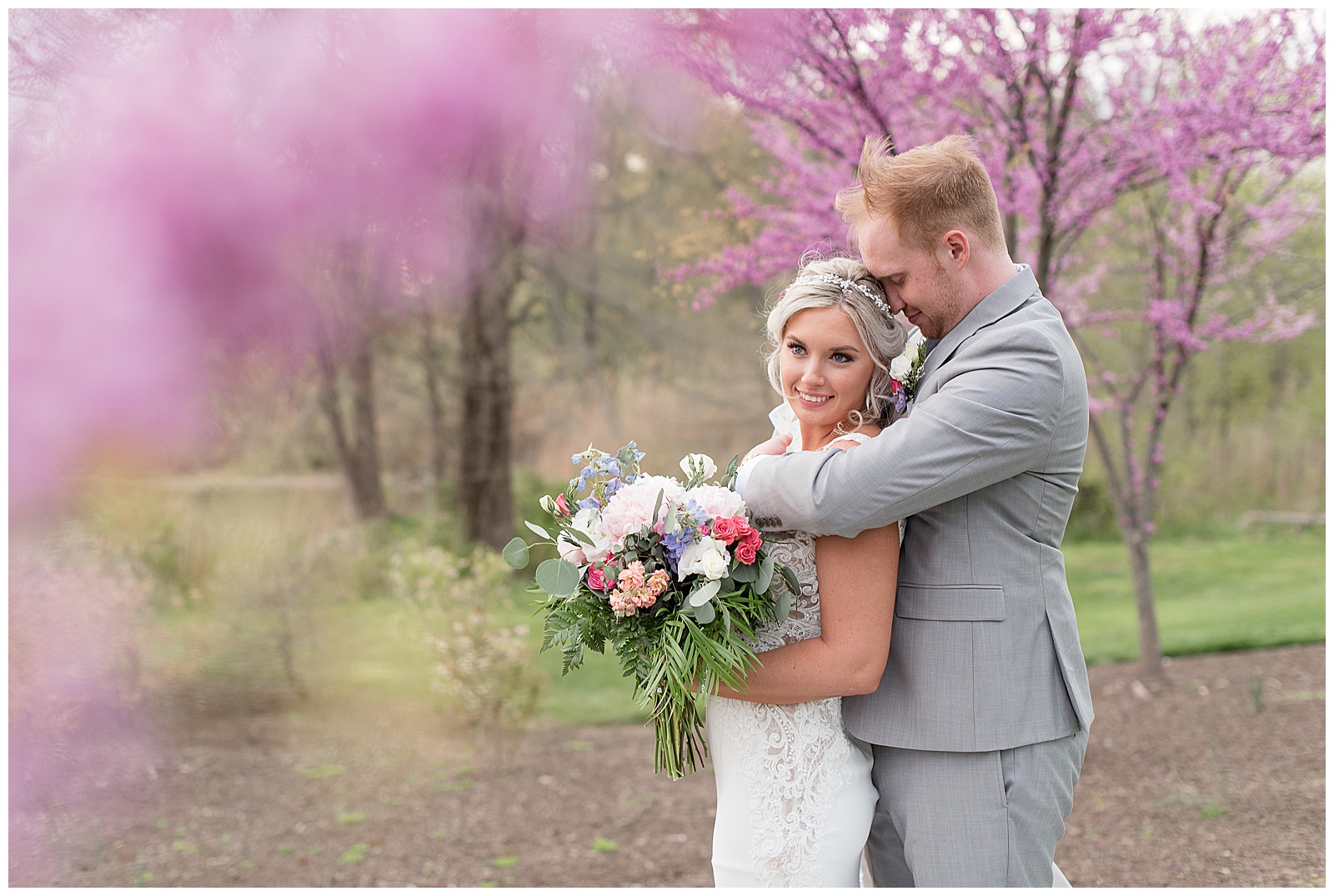 groom standing behind bride hugging her as she smiles at the camera holding bouquet by pink blooming trees