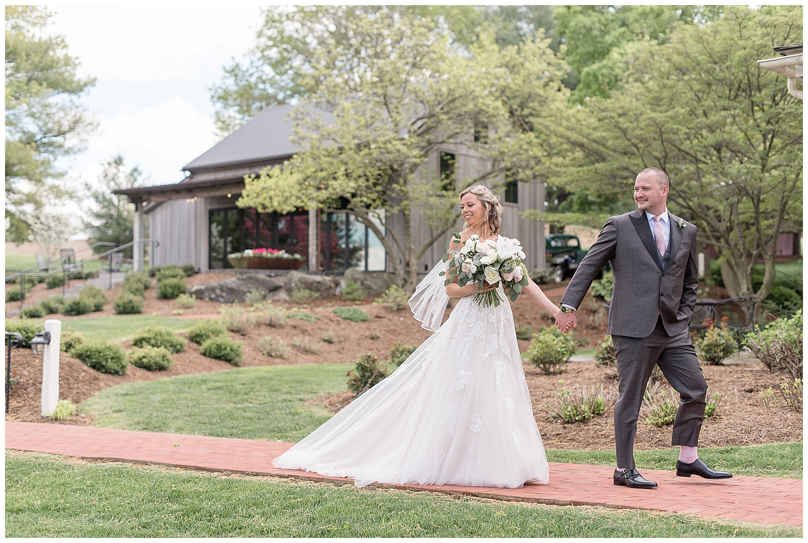 groom leads bride by her hand along brick pathway and both are looking over their right shoulders on spring day at the farm at eagles ridge