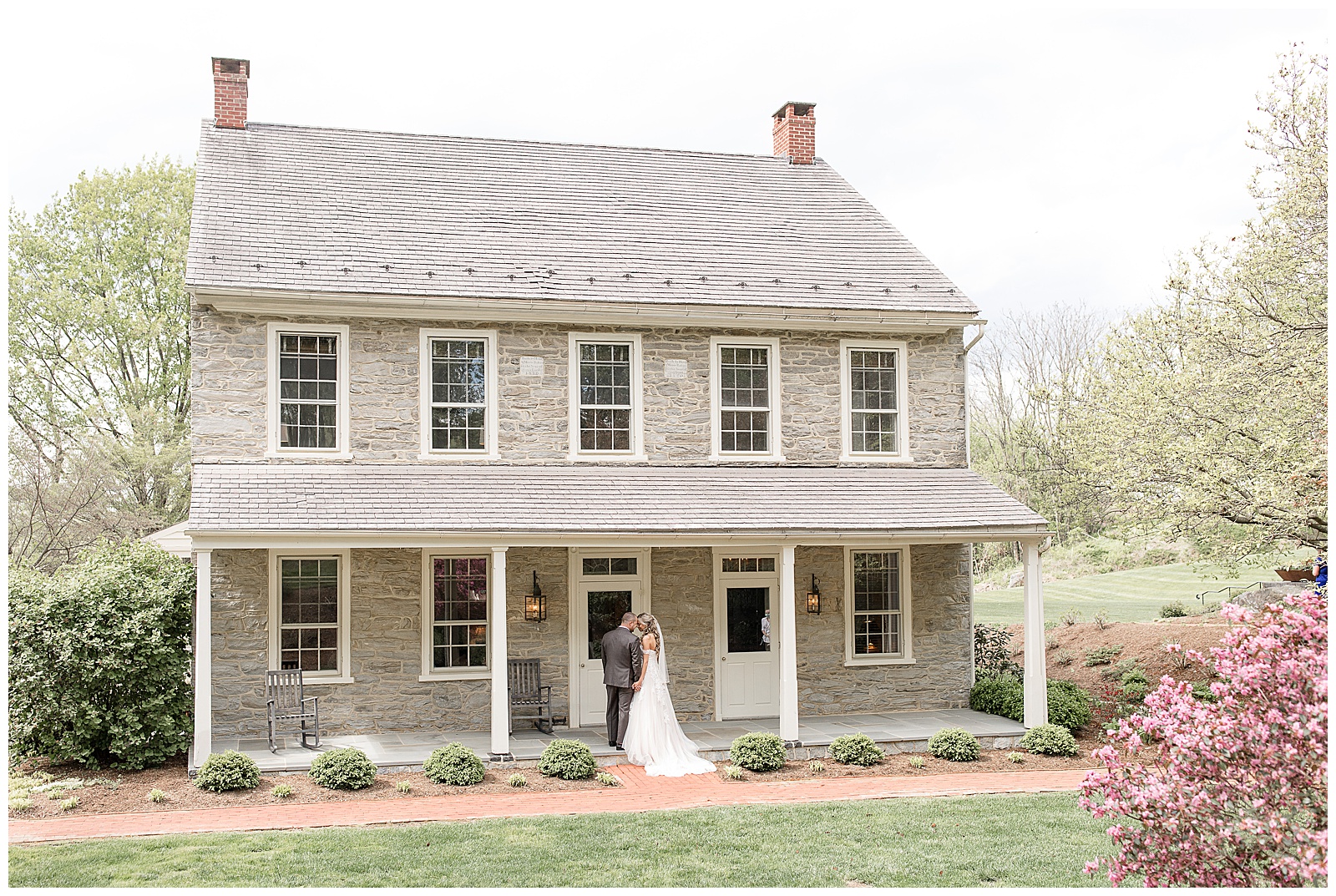 beautiful gray stone farmhouse with bride and groom standing along front edge of porch by landscaped flowerbeds in lancaster pennsylvania