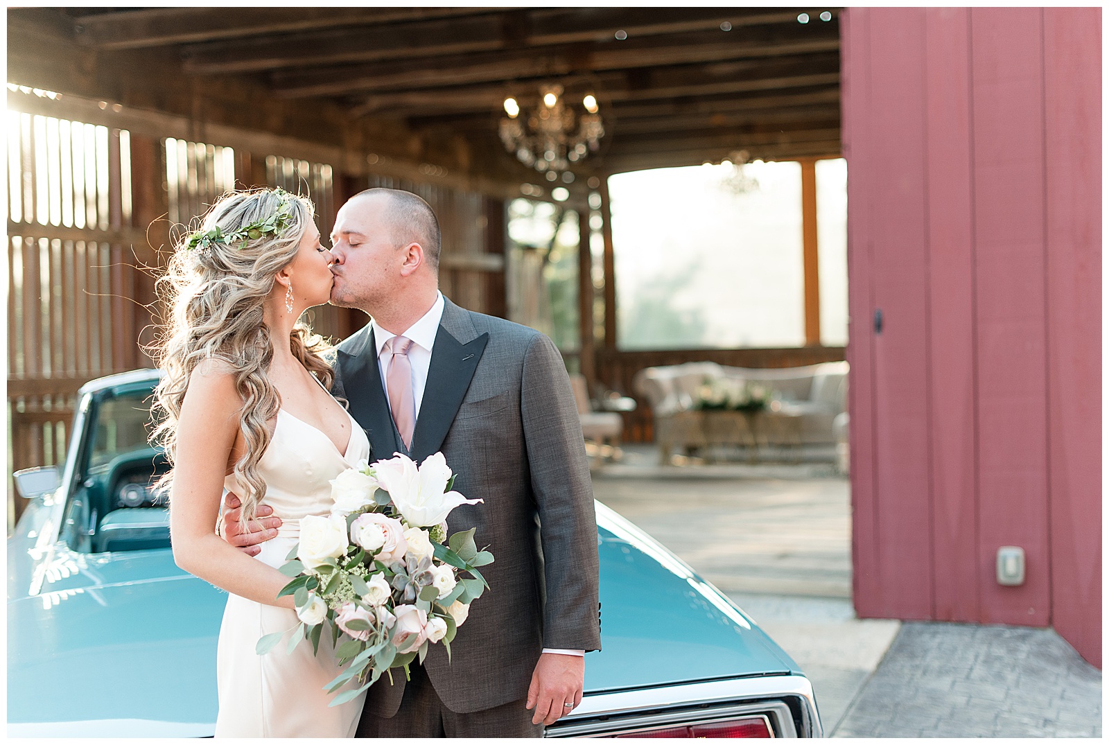 bride and groom kiss while standing behind vintage teal car next to open red barn at the farm at eagles ridge