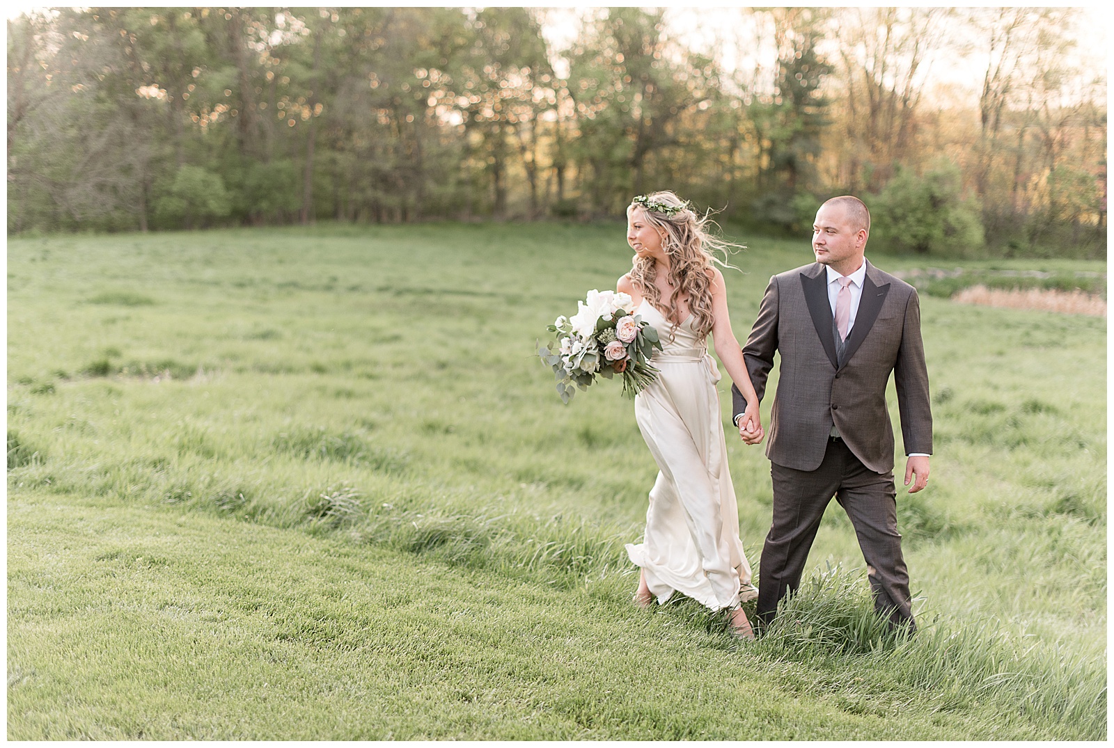 groom leads bride through grassy field as both look back over their right shoulders as sun sets at the farm at eagles ridge
