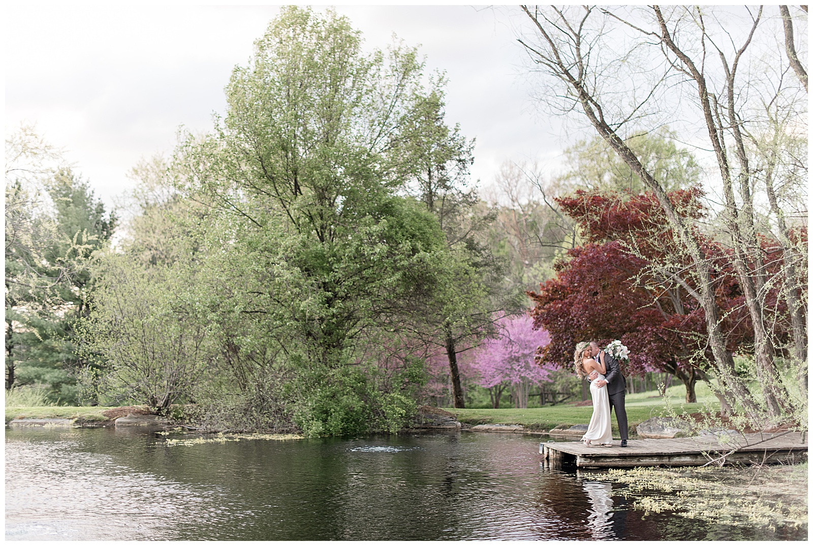couple kissing at the end of wooden dock along pond and surrounded by blooming trees on sunny day in lancaster pennsylvania