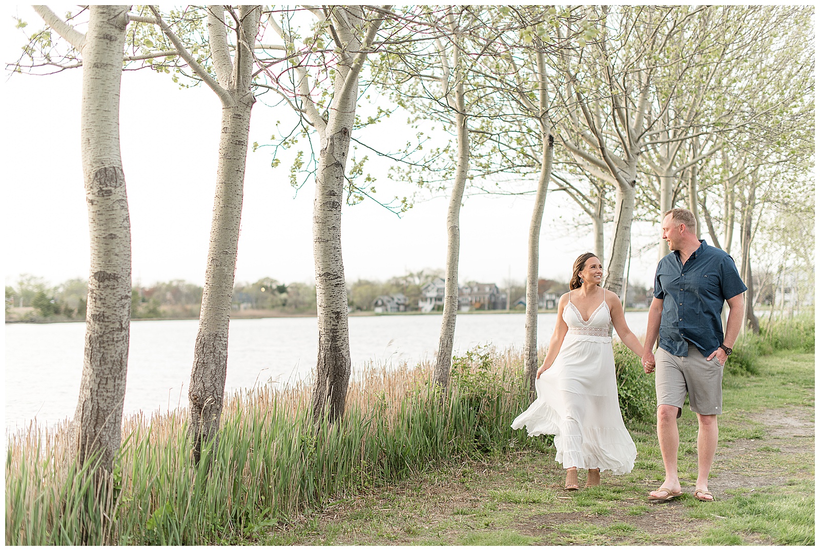 couple walking along bay head harbor tall grasses and row of trees holding hands and smiling
