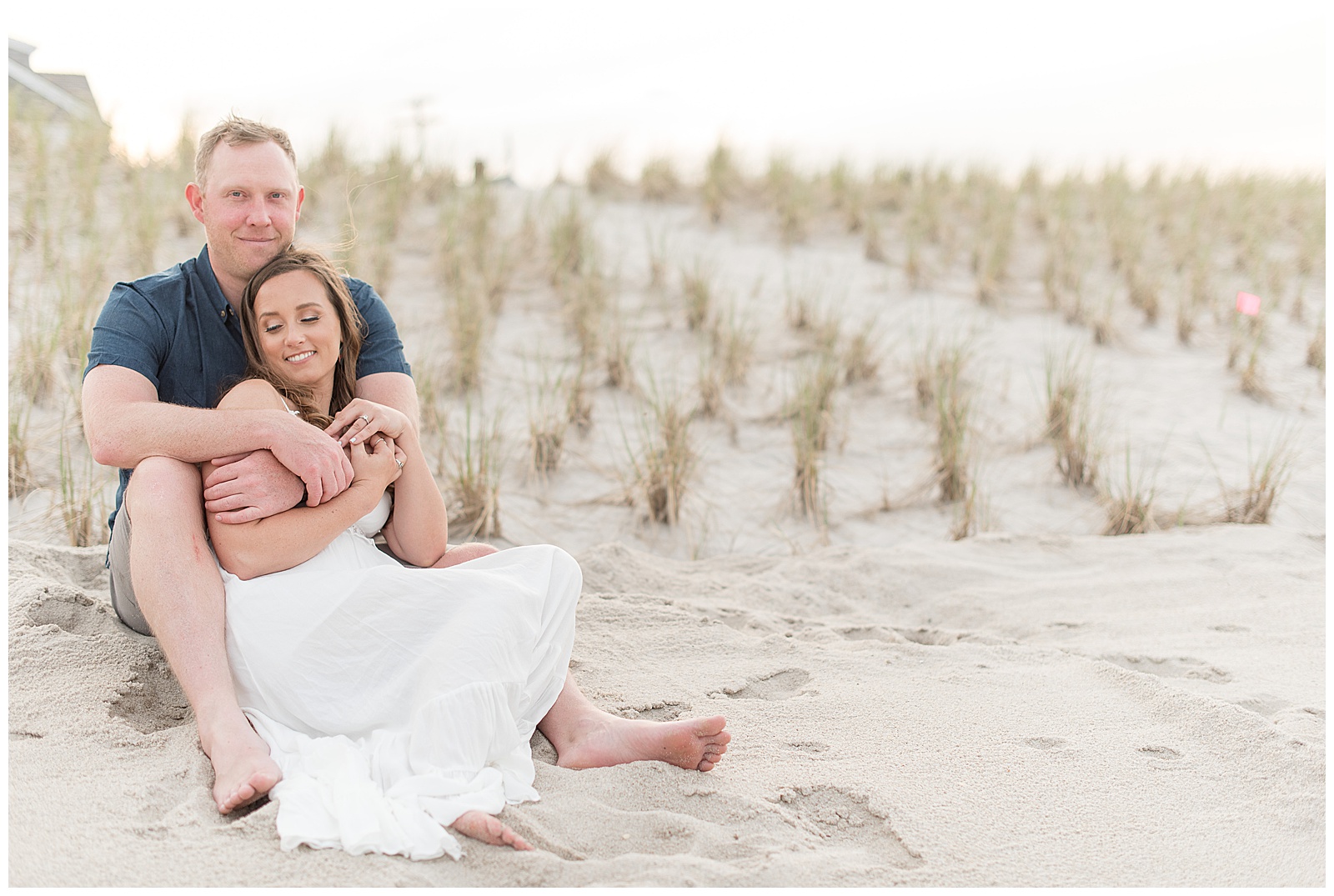 woman snuggles in tightly by man as they sit in the sandy dunes of bay head new jersey on a sunny evening