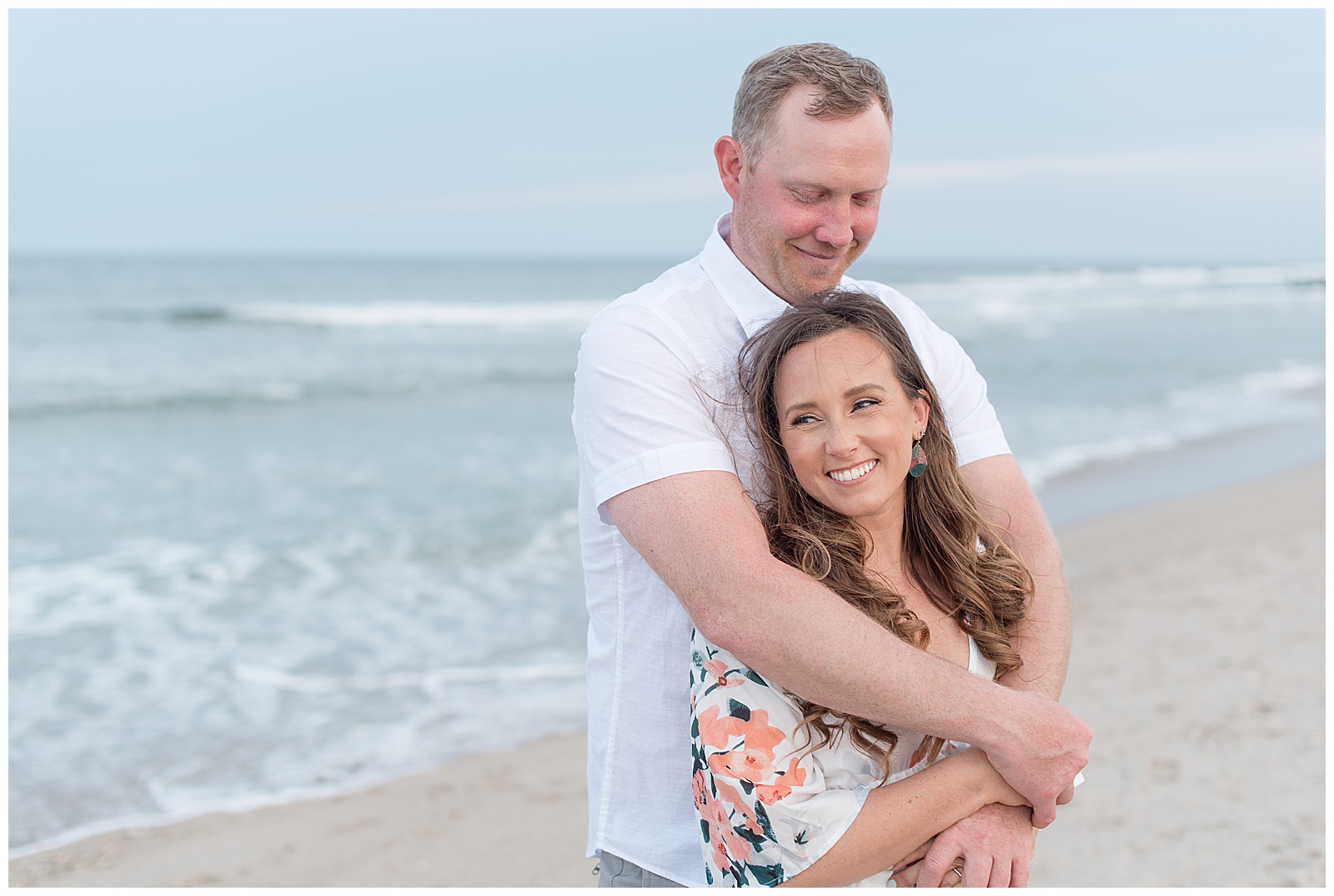 guy hugs girl from behind as they both smile looking away from camera along the water in bay head