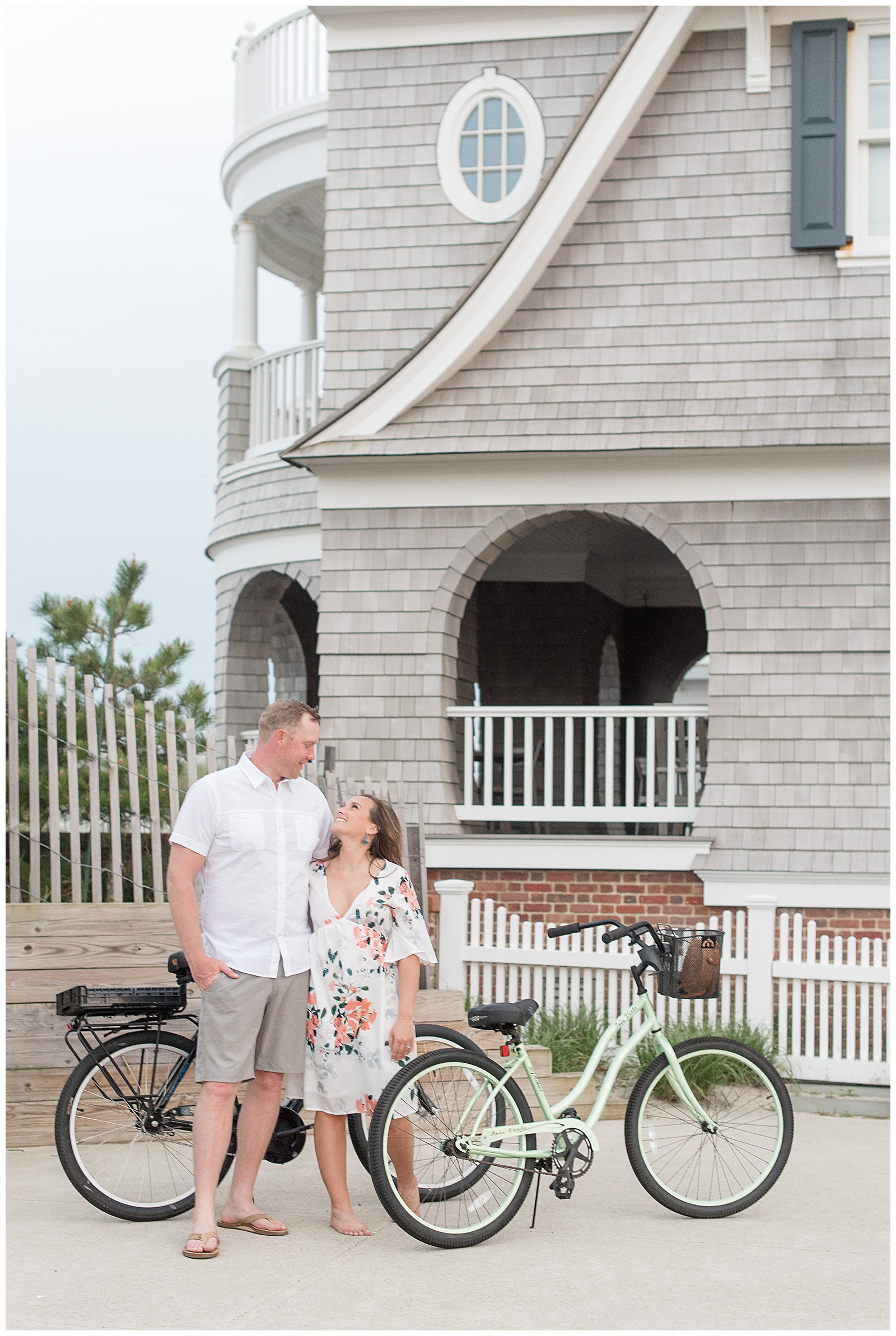 couple looking at one another smiling next to two beach bikes and a beautiful gray beach house in new jersey