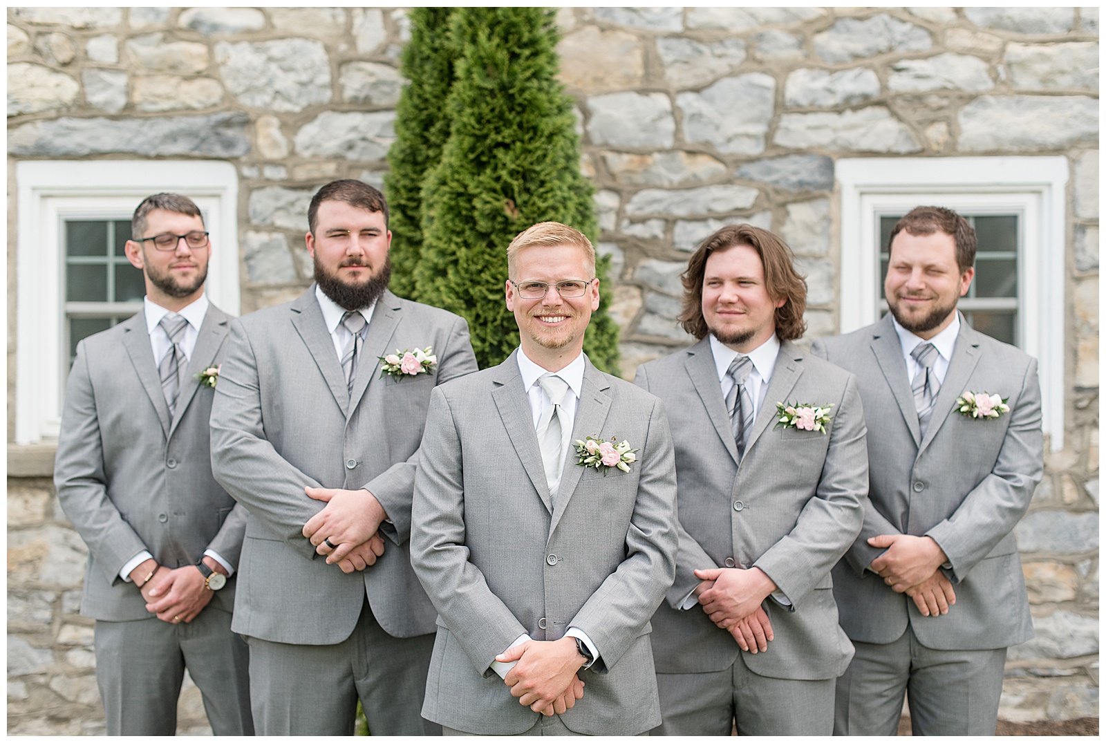 groom and his groomsmen have their hands folded in front of themselves and are smiling at the camera