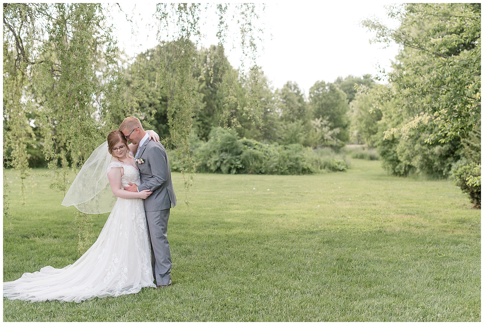 bride looks down as groom kisses the left side of her forehead in grass lawn by willow tree at the barn at silverstone