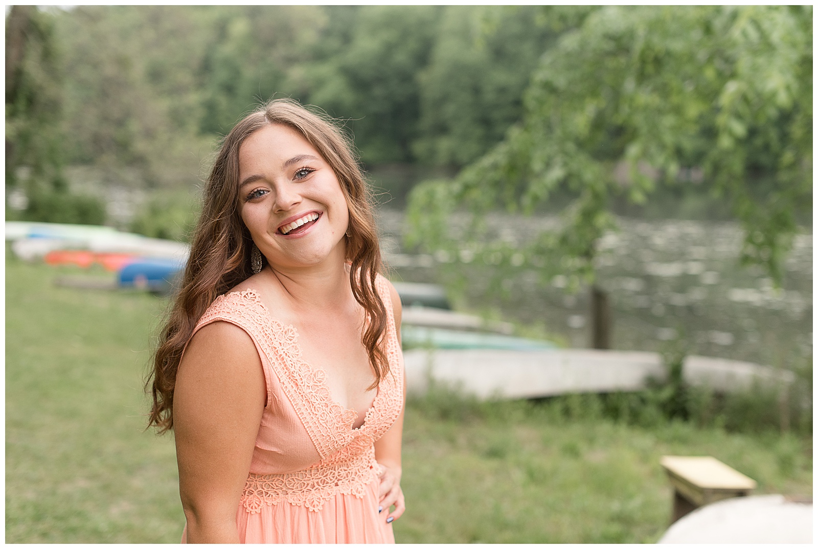 senior girl smiling at camera with left hand on hip and head tilted back near lake in lititz pennsylvania