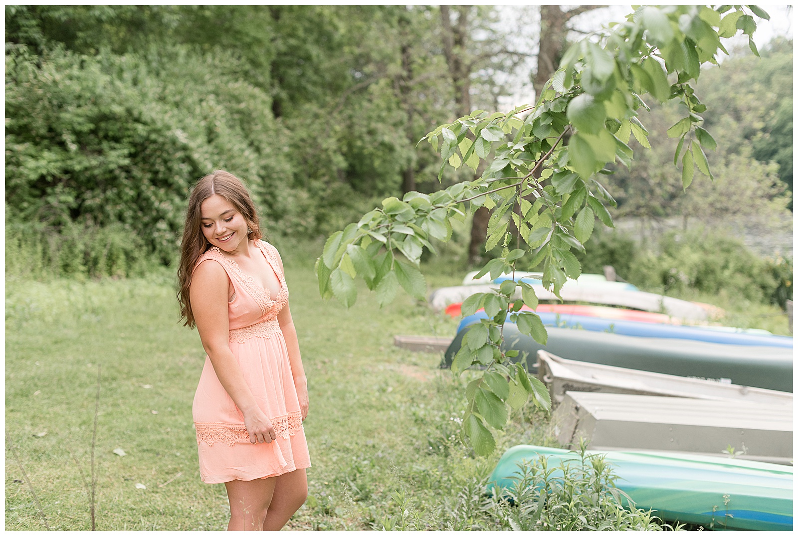 senior girl facing colorful canoes holding edge of dress while looking down and smiling in lititz pennsylvania