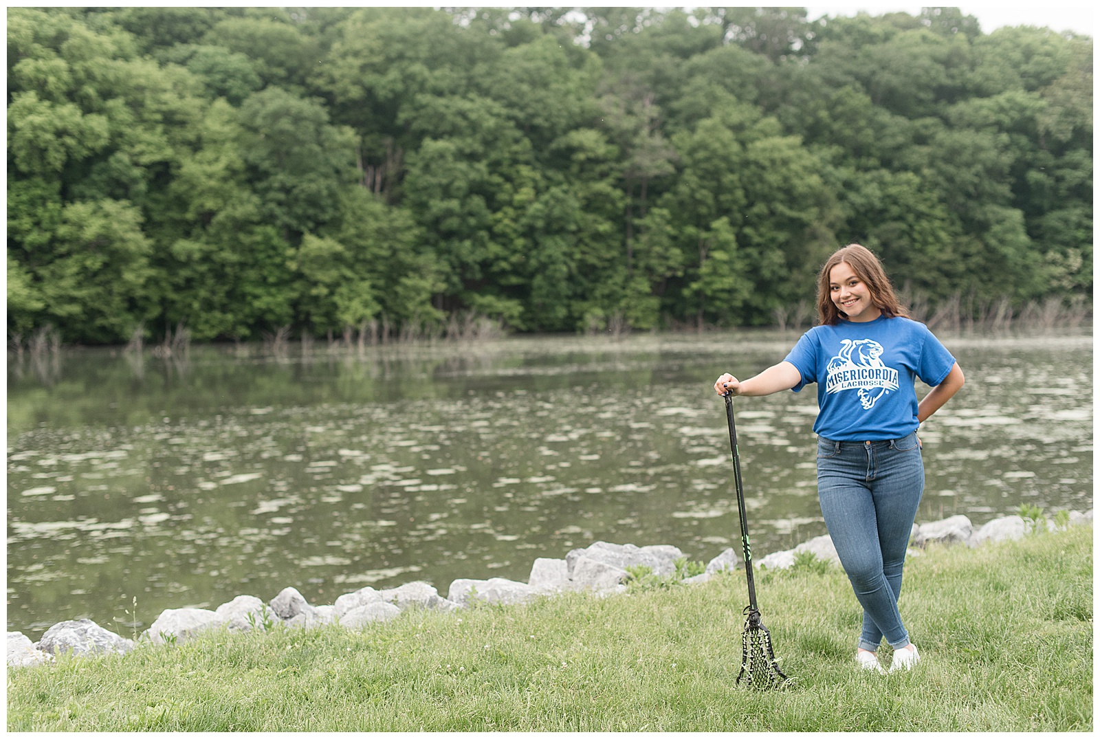 senior girl wearing lacrosse t-shirt and blue jeans leaning on black lacrosse stick along speedwell forge lake in lititz pennsylvania