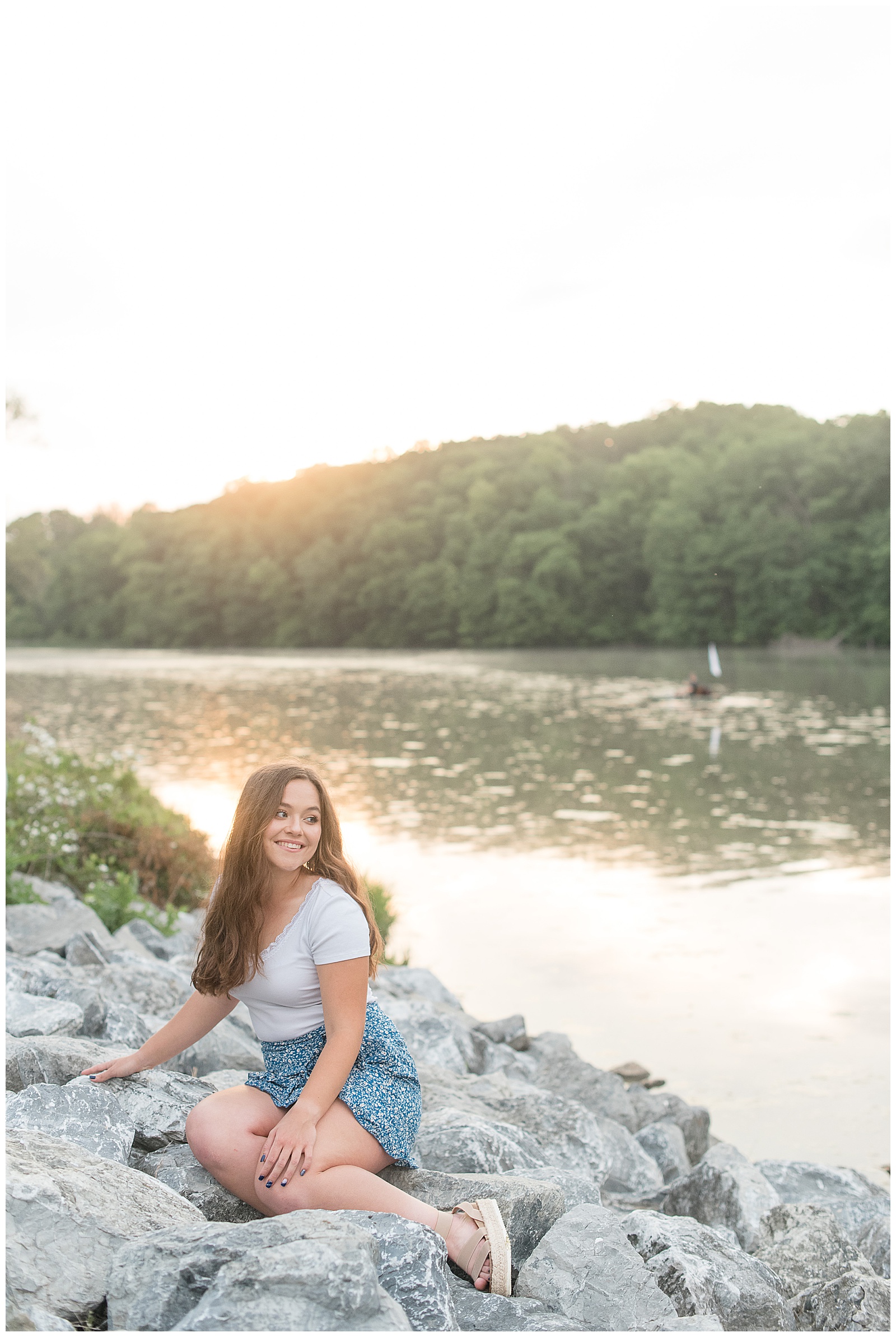senior girl sitting on large rocks looking out over speedwell forge lake near sunset in lititz pennsylvania