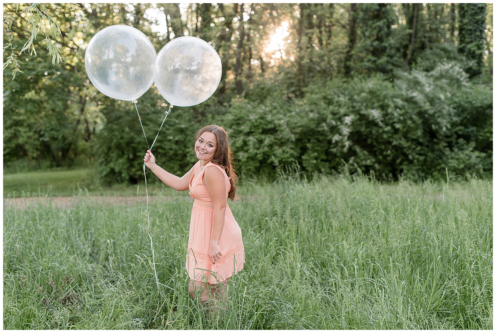 senior girl in peach colored dress holding two large clear balloons standing in tall grass at overlook park