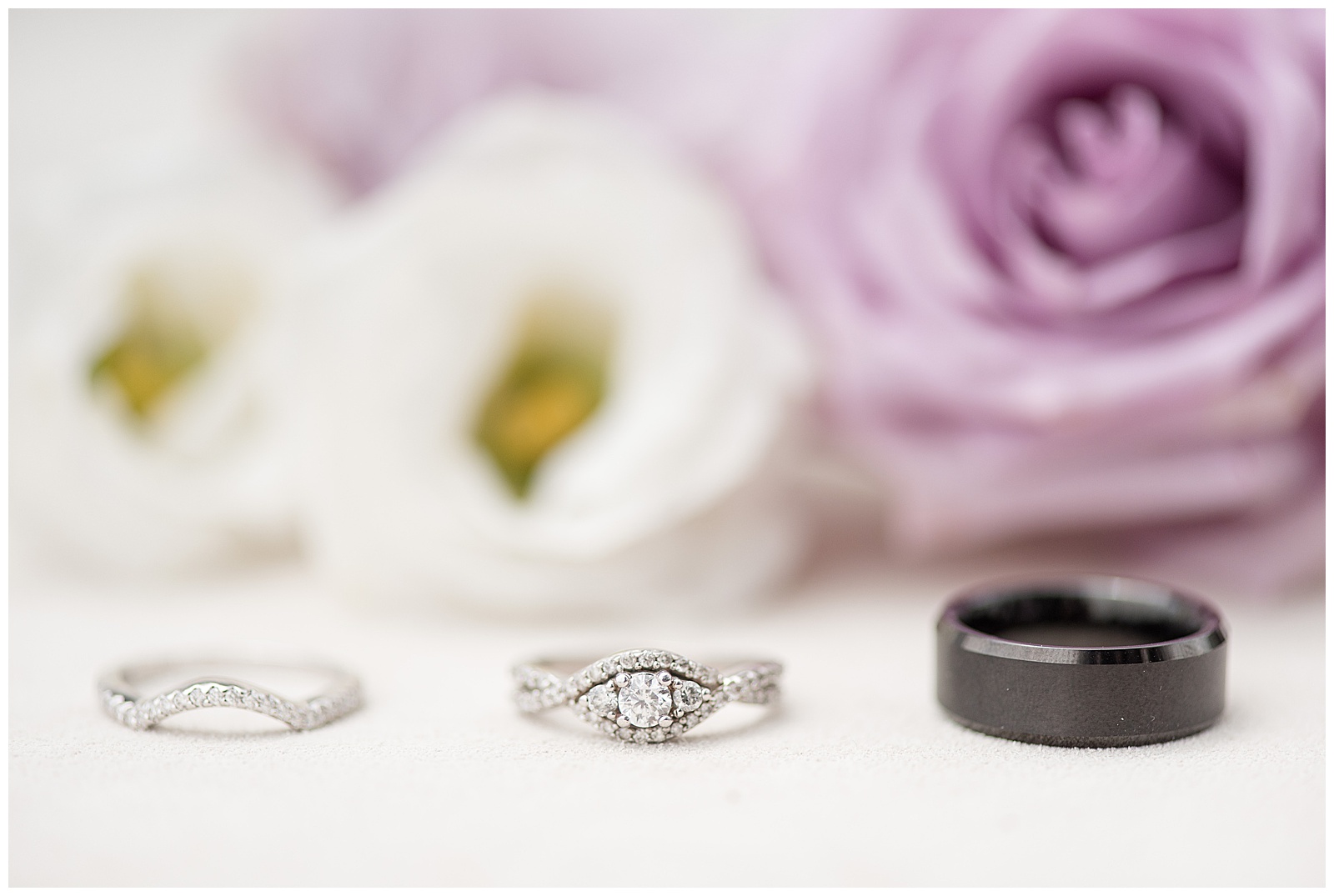 silver and diamond wedding ring and engagement next to groom's black wedding band with white and lavender roses behind