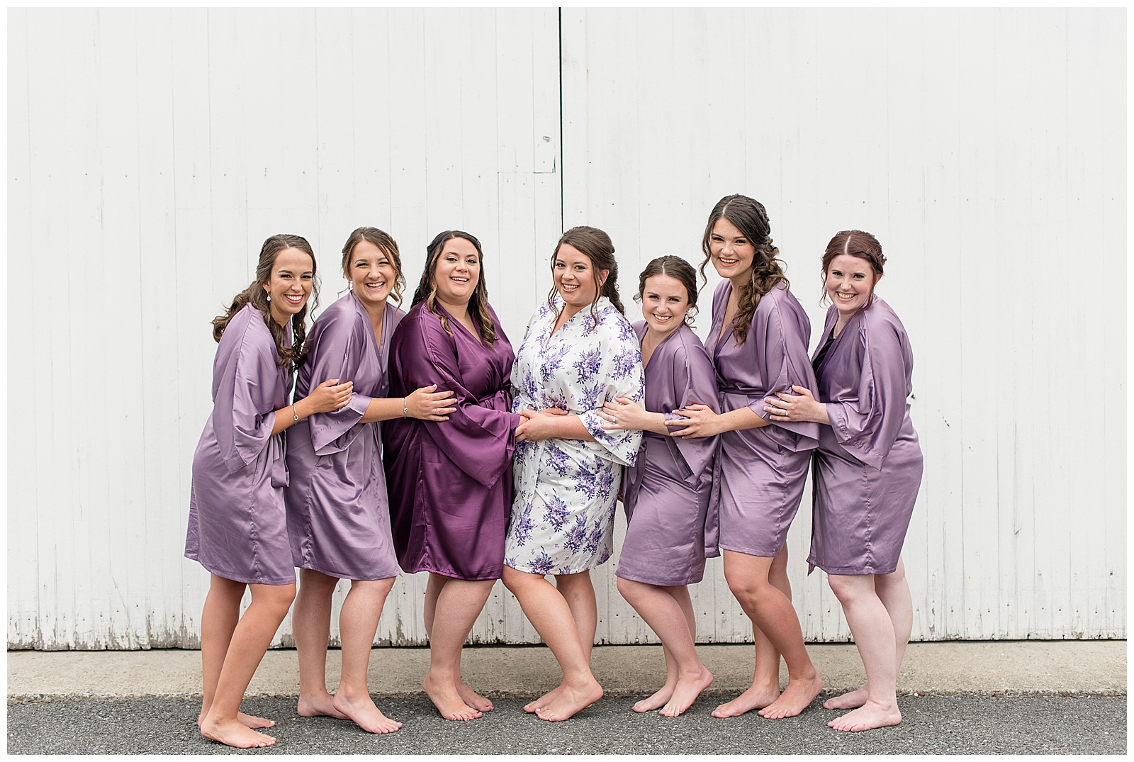 bride and bridesmaids in floral and lavender silk robes standing close with arms touching by white barn door in lancaster, pennsylvania