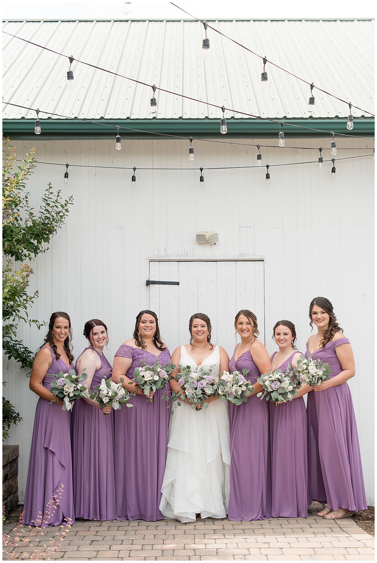 bride and bridesmaids standing close holding white and lavender bouquets outside white barn in manheim, pennsylvania