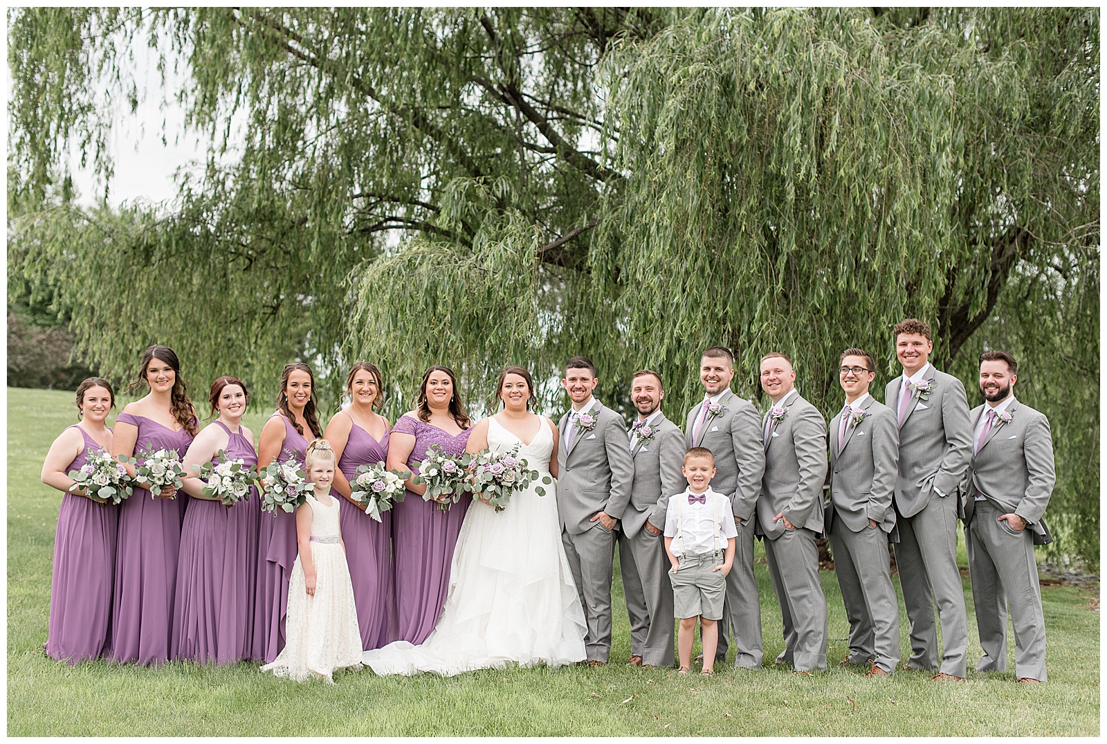 couple surrounded by wedding party in lavender dresses and light gray suits under willow tree at lakefield weddings