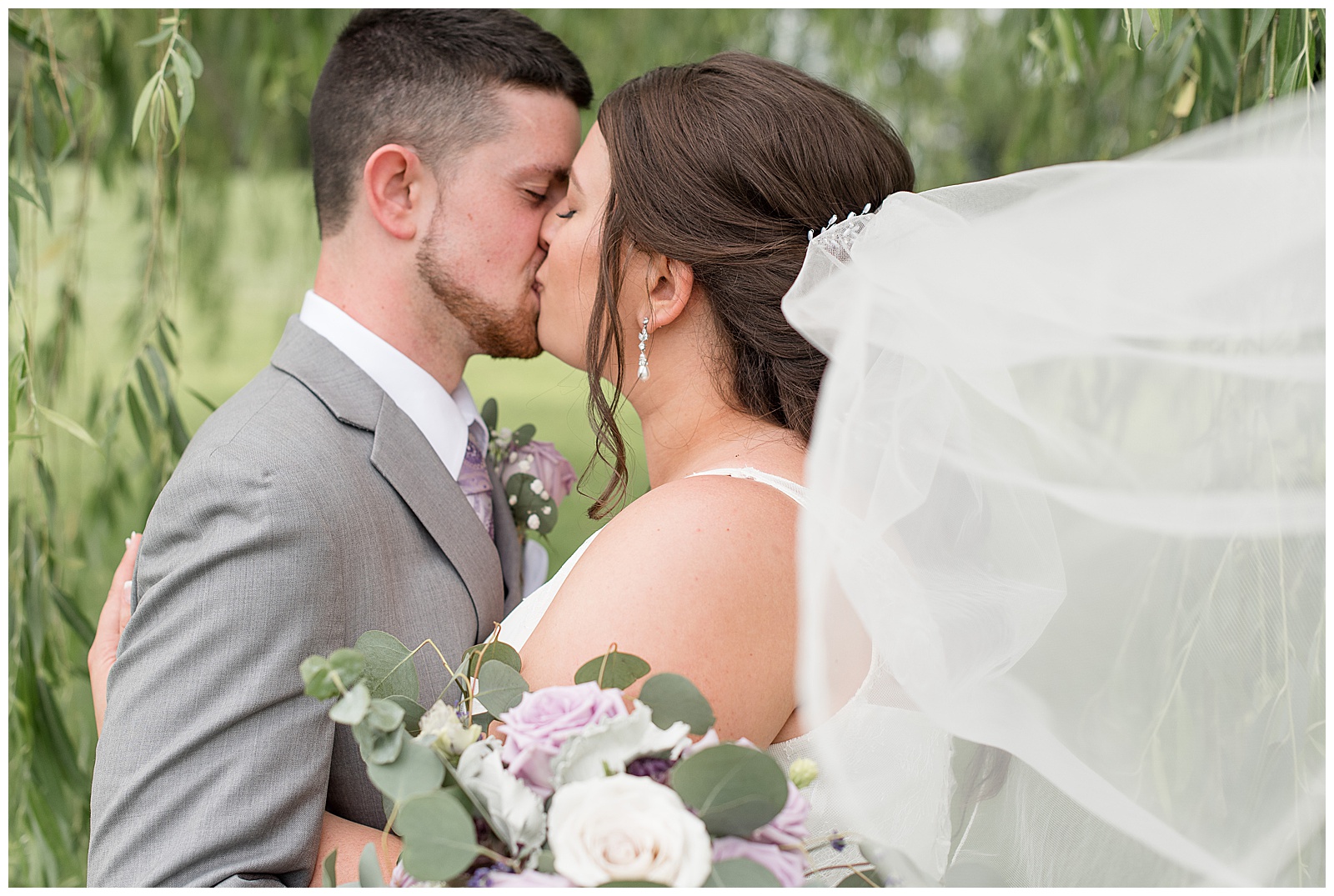 close up of groom kissing bride as her veil blows in the wind and he holds her bouquet at lakefield weddings