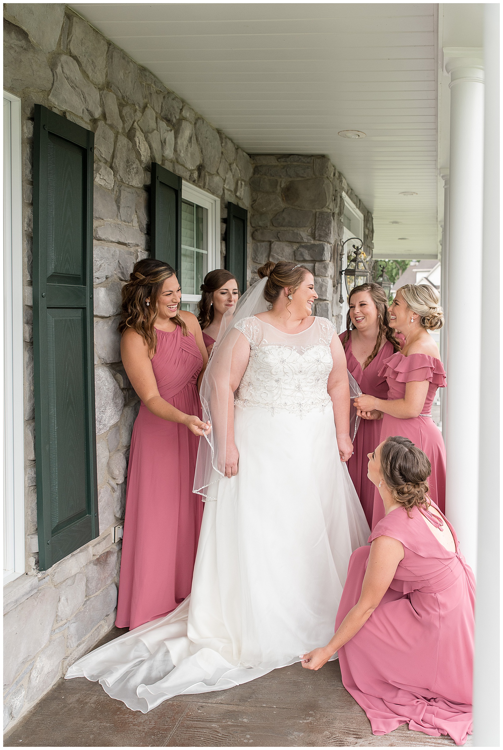 bride glances over left shoulder smiling at her bridesmaids surrounding her wearing blush gowns on front porch in lancaster pennsylvania