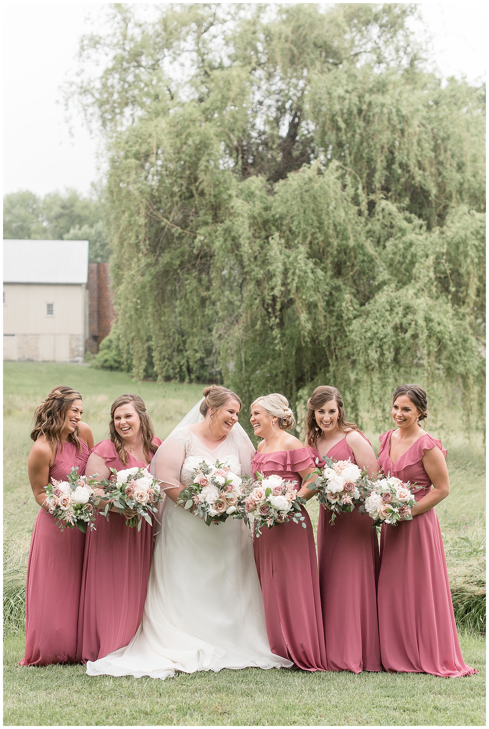 bride and bridesmaids all looking at each other and smiling holding bouquets near willow tree in strasburg pennsylvania