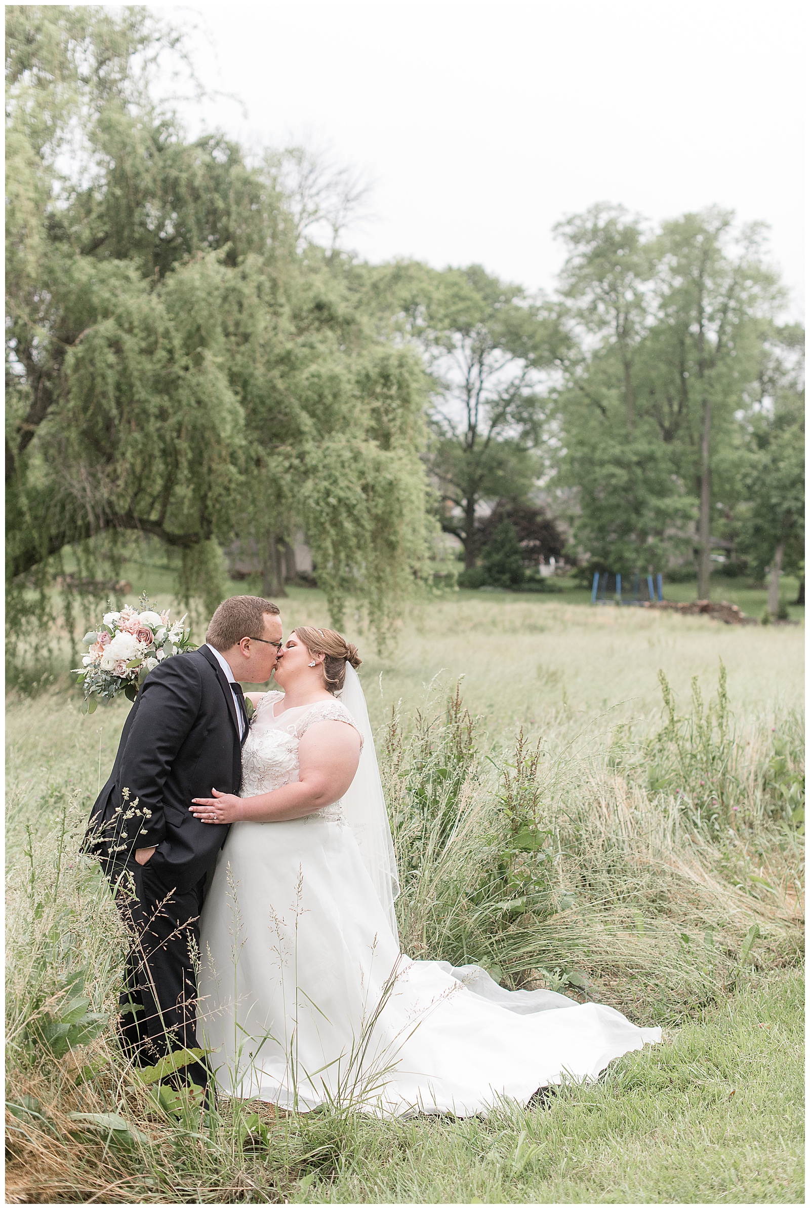 groom kisses bride as they hug in tall grasses near willow tree at willows at historic strasburg wedding venue