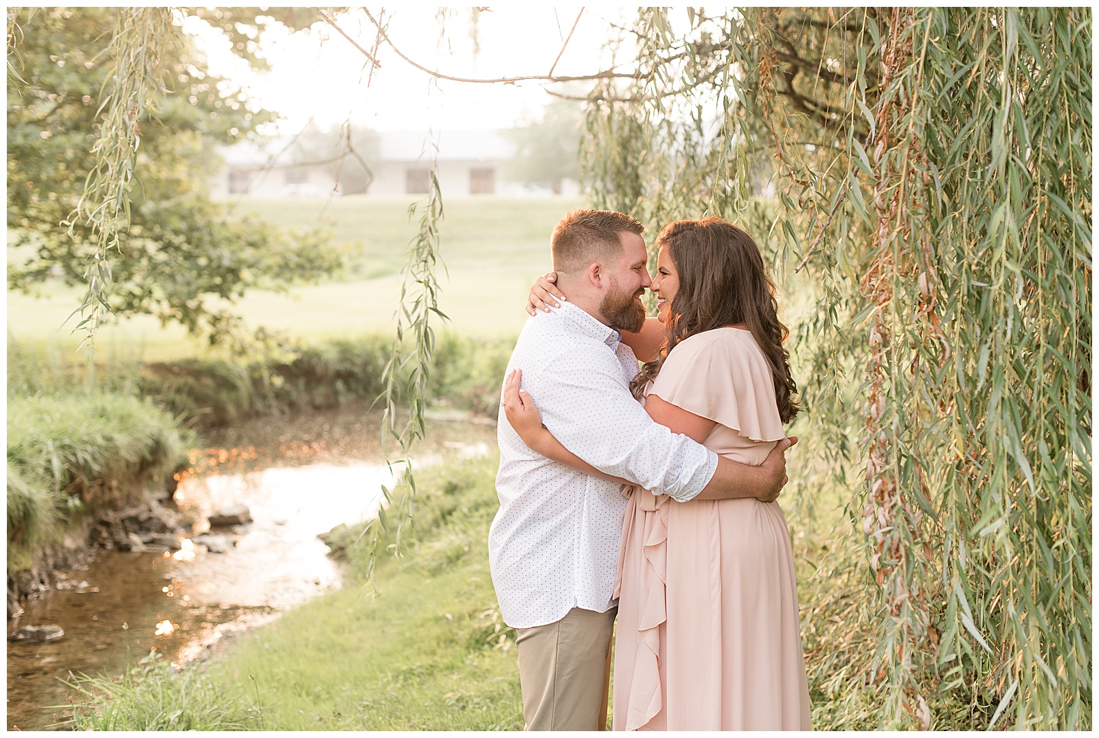 couple hugging and almost kissing under willow tree beside stream with sun shining behind them in lancaster pennsylvania