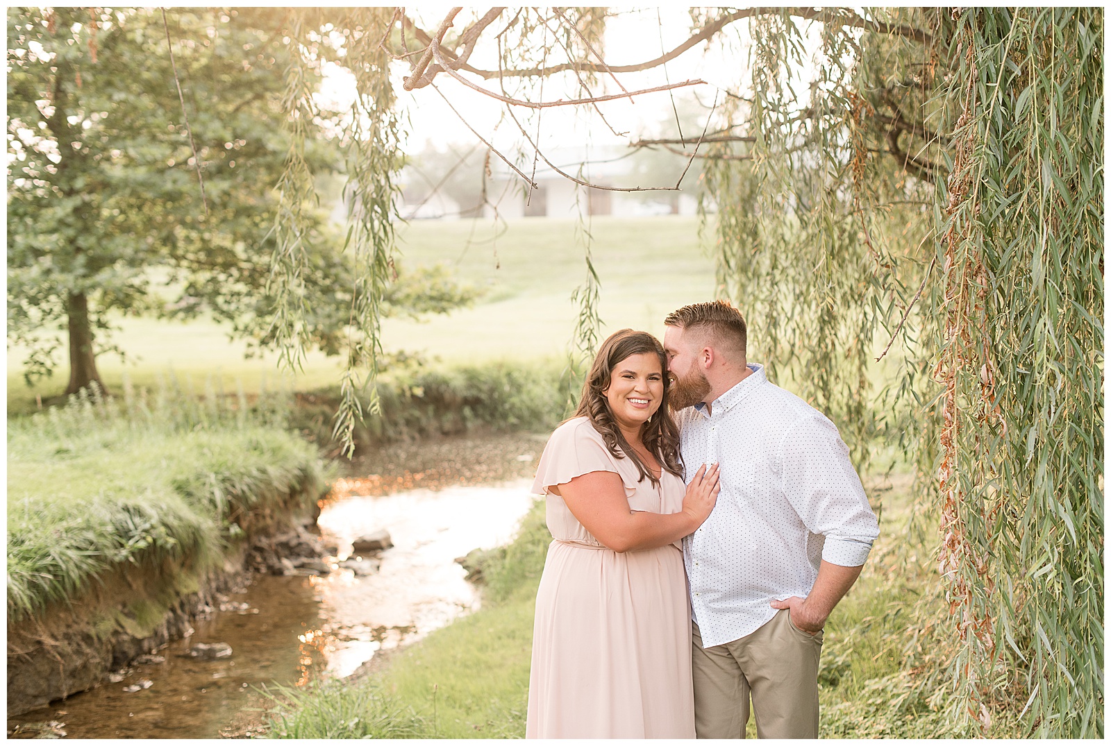 guy kissing girl as she smiles at camera and rests her right hand on his chest next to small stream under willow tree in lancaster pennsylvania