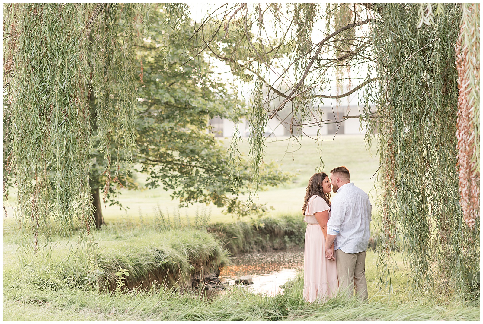 couple holding hands and kissing along small stream under willow tree on sunny evening at conestoga house and gardens