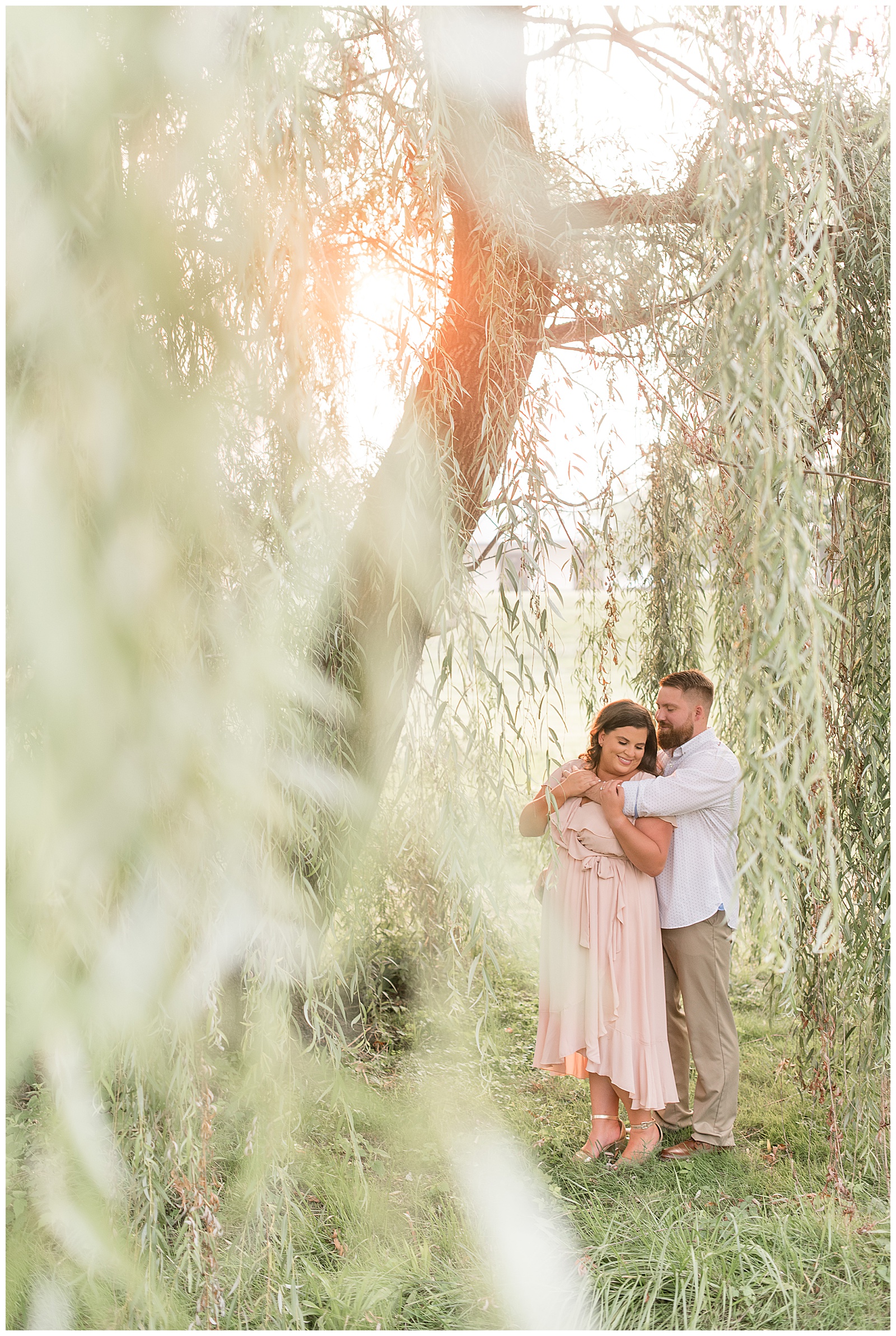 guy hugging girl from behind as she looks down and standing under large willow tree and sun shining in lancaster pennsylvania