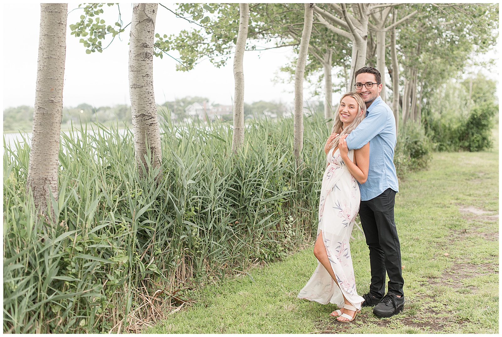 guy hugs girl from behind as they both smile looking at the camera next to tall bay grasses in new jersey