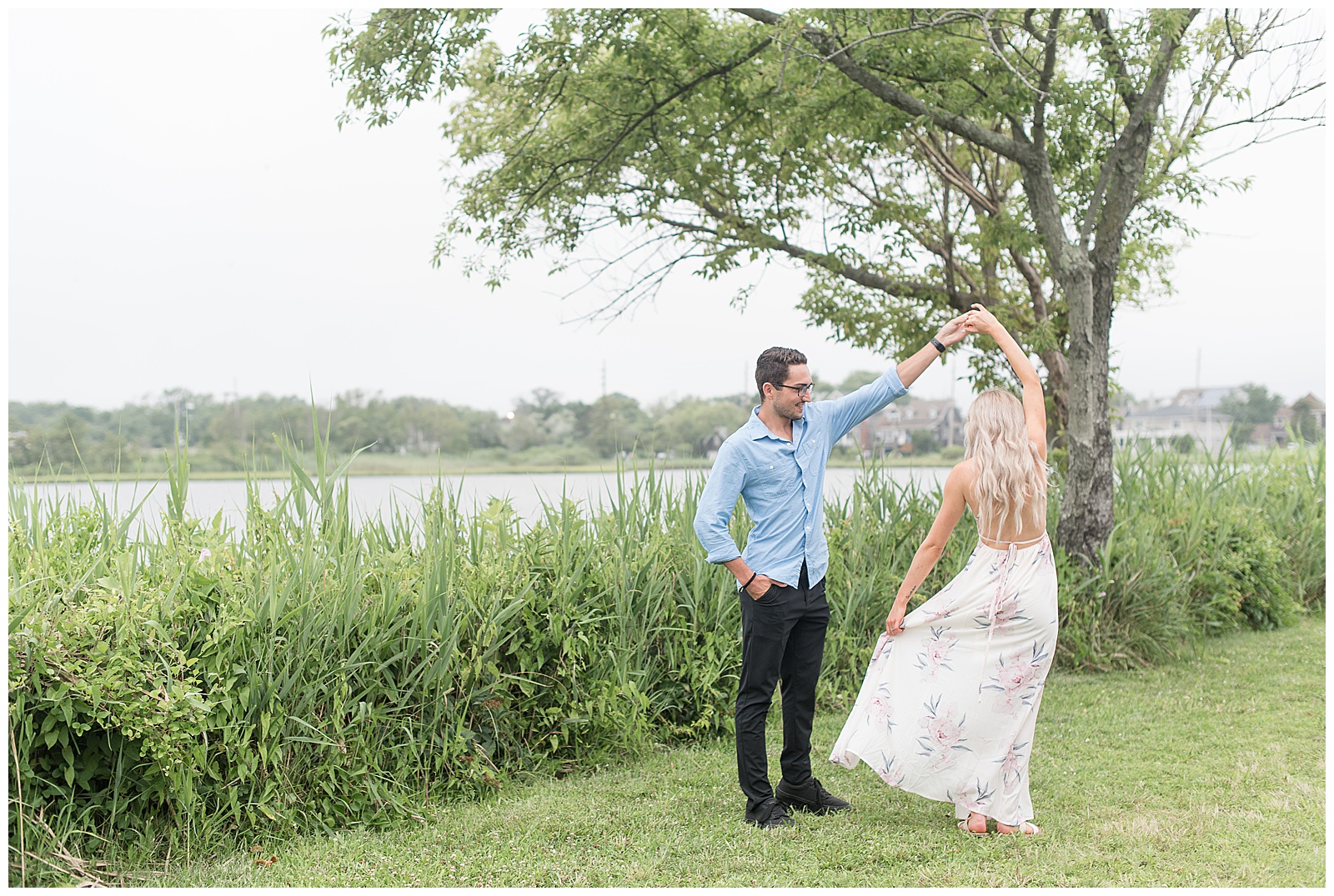 guy twirls girl under his left arm as she flares her flowy dress along plants by the bay