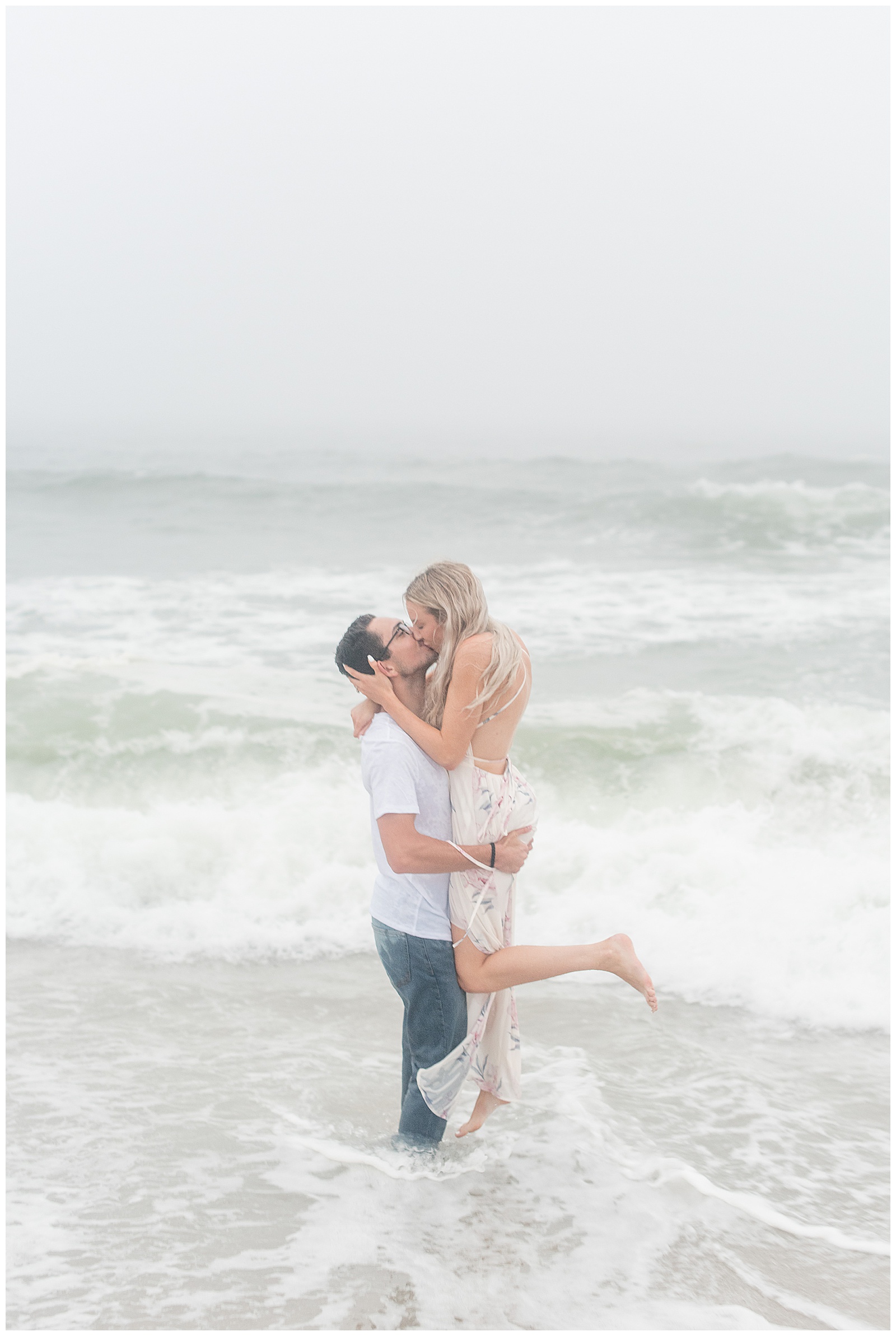 guy in jeans and white t-shirt stands in ocean holding and kissing girl in floral dress