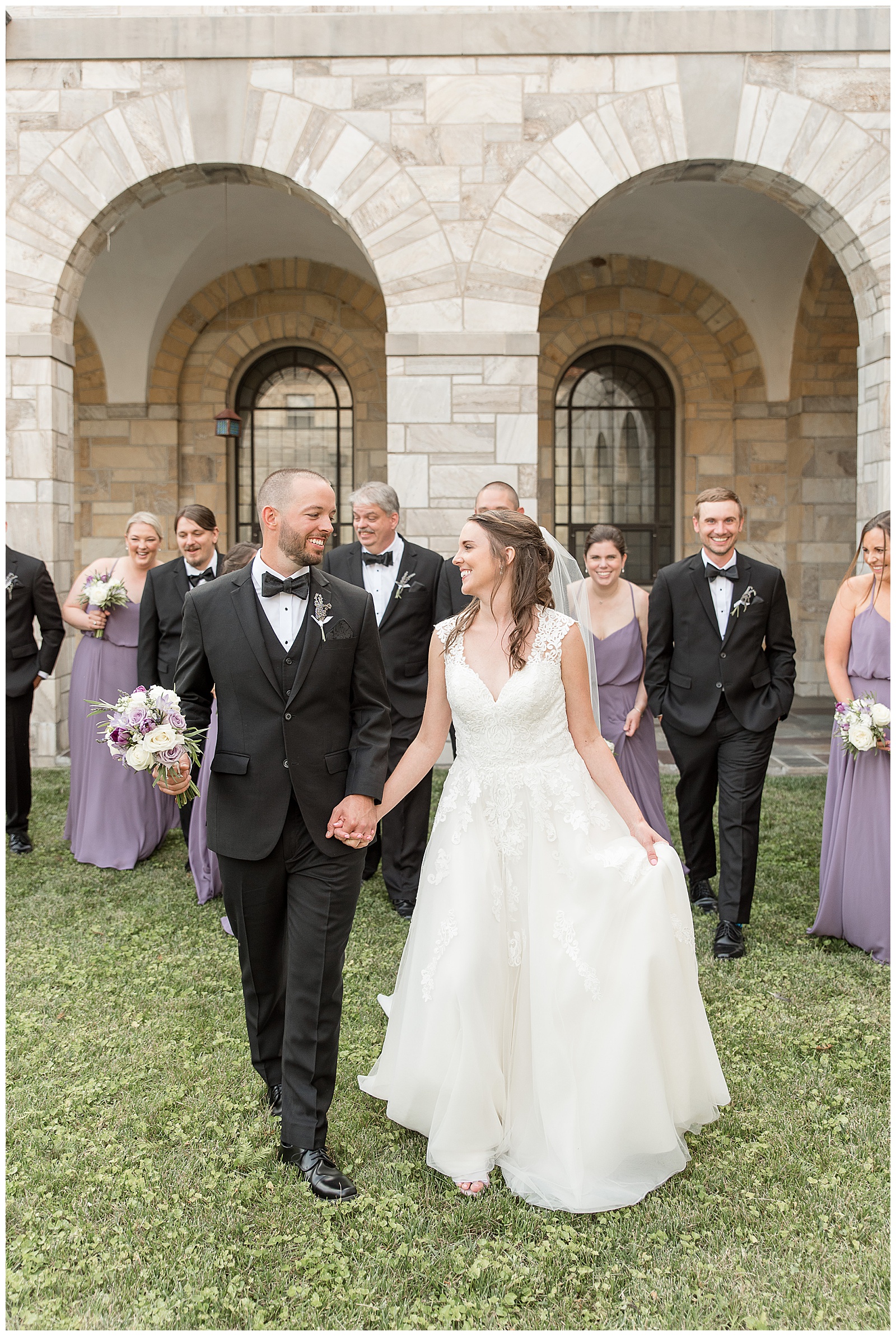 bride and groom walking hand-in-hand smiling at each other with bridal party walking behind