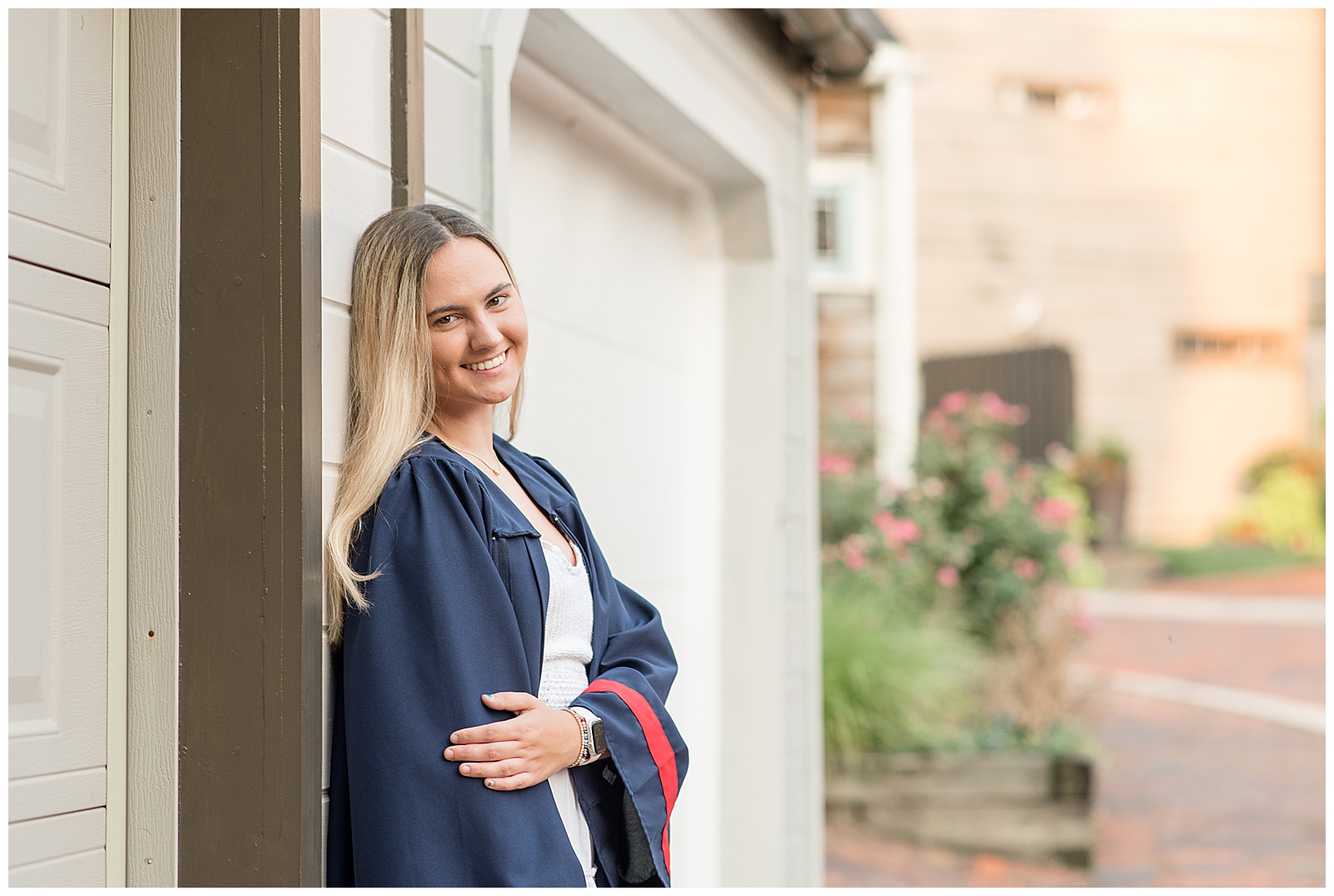 senior girl in navy blue graduation gown leans against tan building smiling with her arms folded around her stomach in lancaster city
