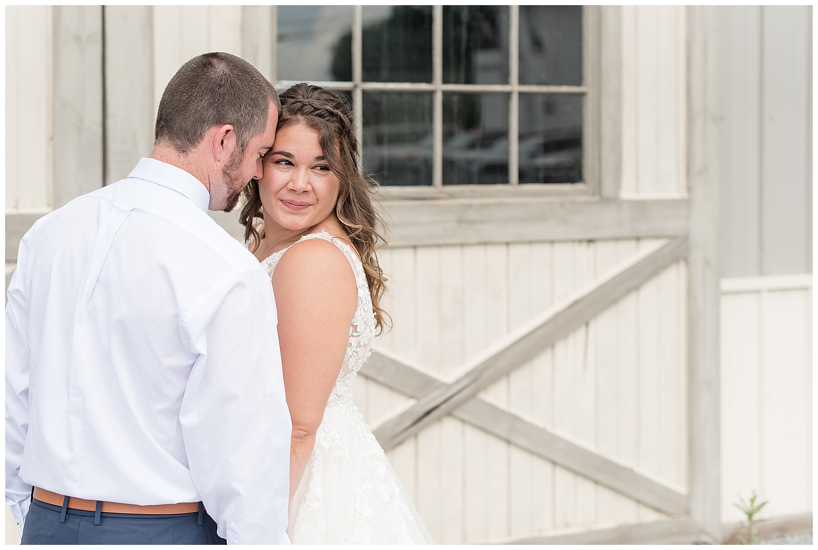 bride looking off camera as groom rests head on her temple in front of white barn doors