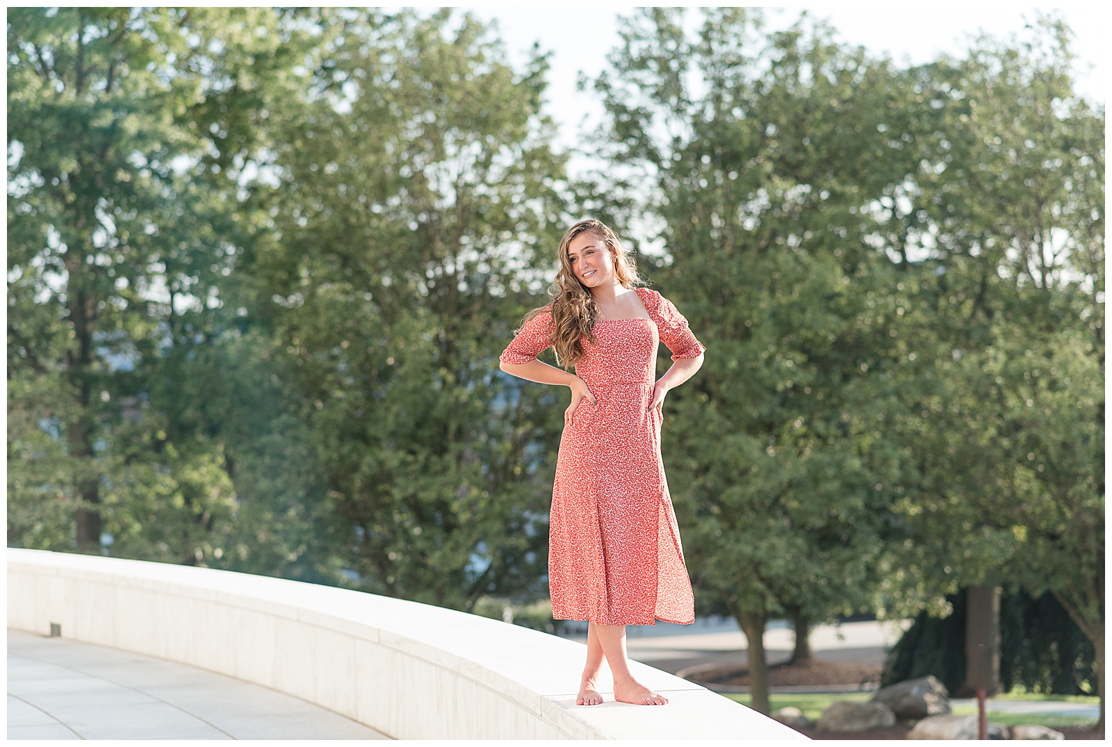 senior girl in red floral dress standing atop concrete structure with hands on hips and trees behind her