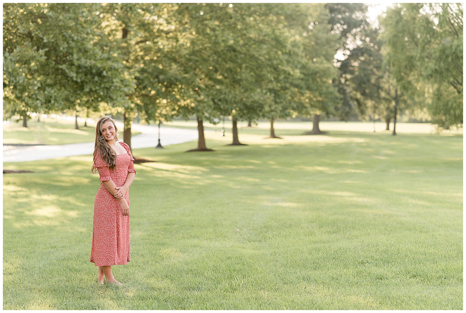 senior girl in long red floral dress standing in grass lawn with row of trees behind her at founders hall